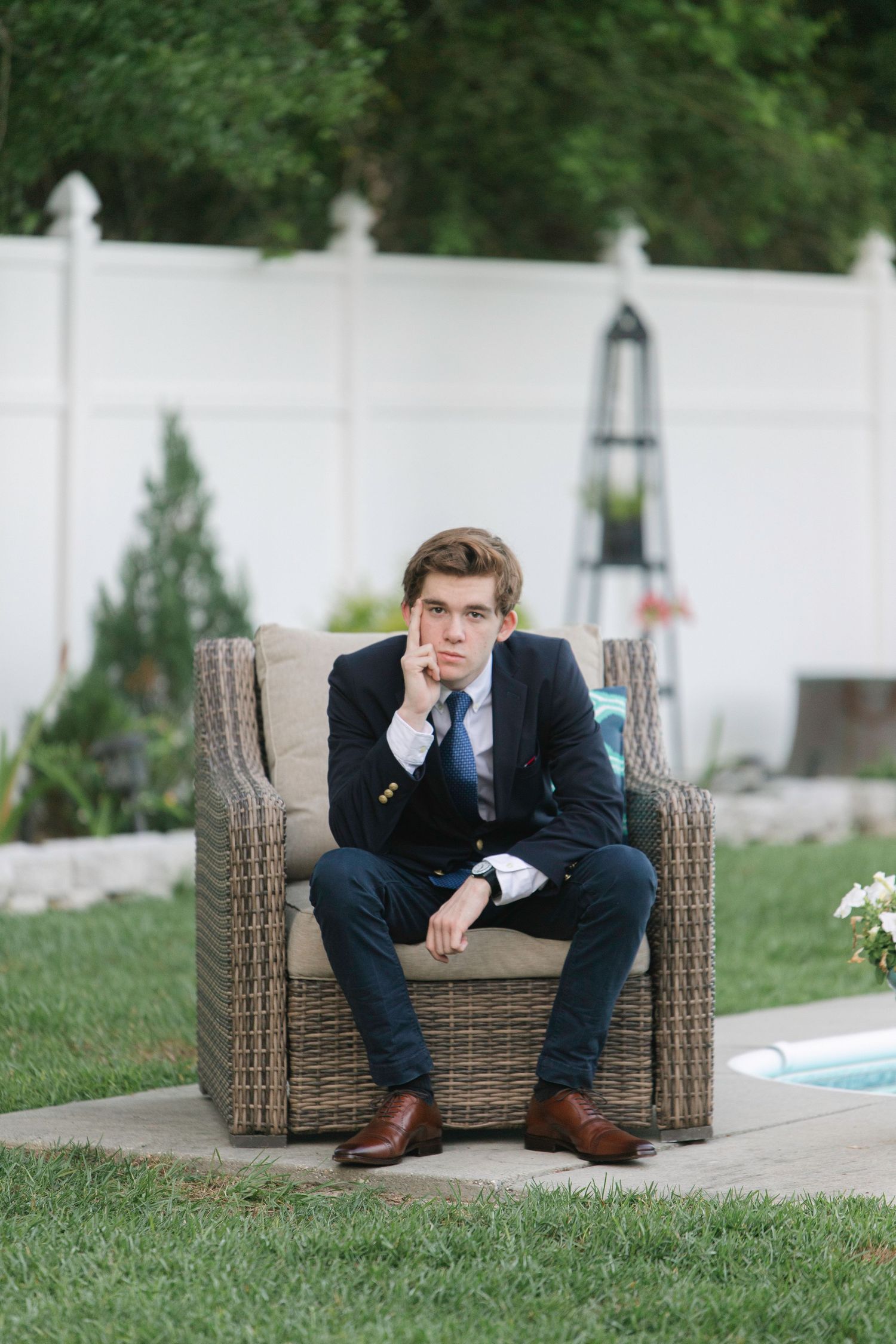 Young man in navy suit sitting on wicker chair in backyard garden setting, looking thoughtful.