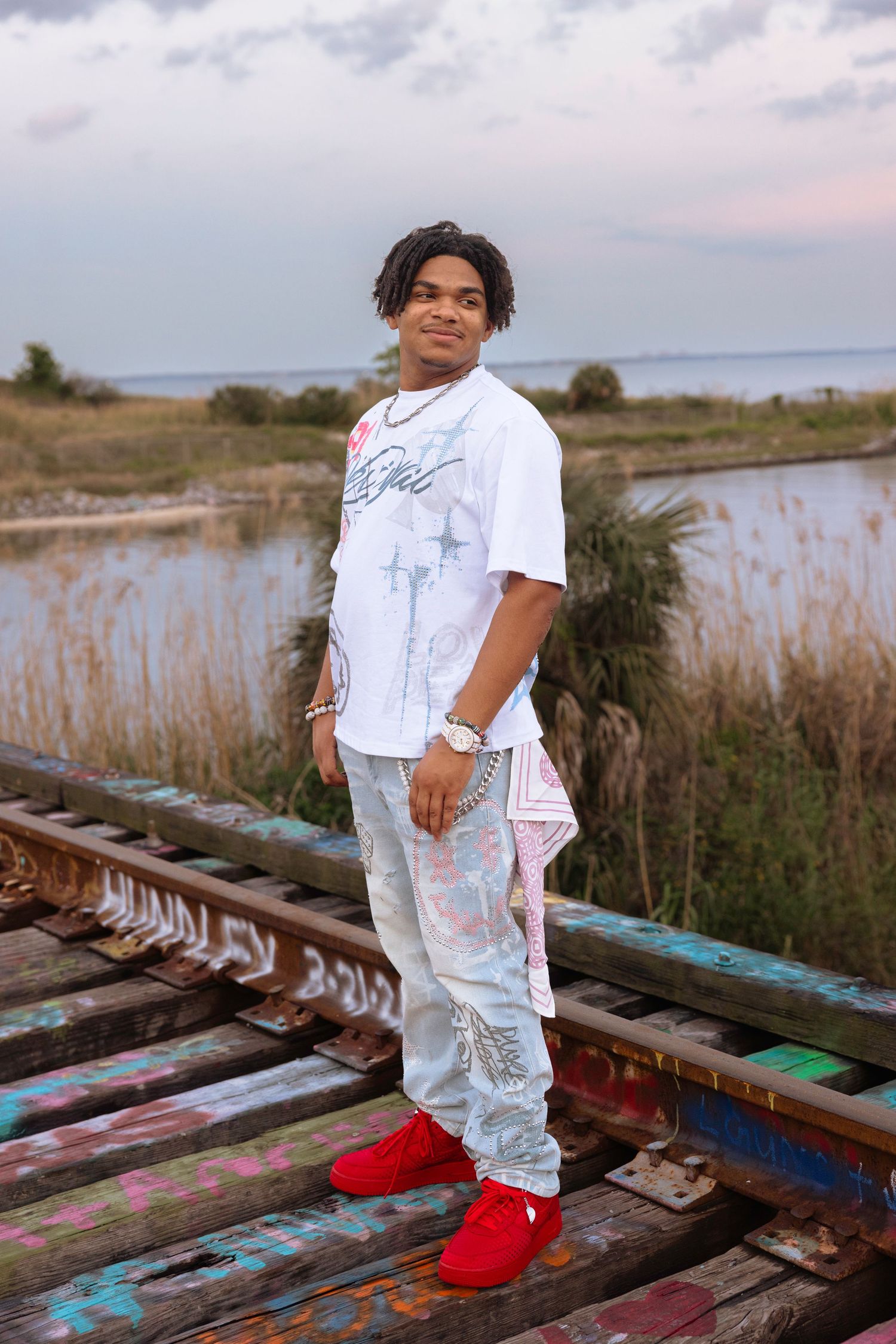 Young man in white graphic tee, light jeans, and red sneakers posing on a colorful graffiti bridge near a marsh.