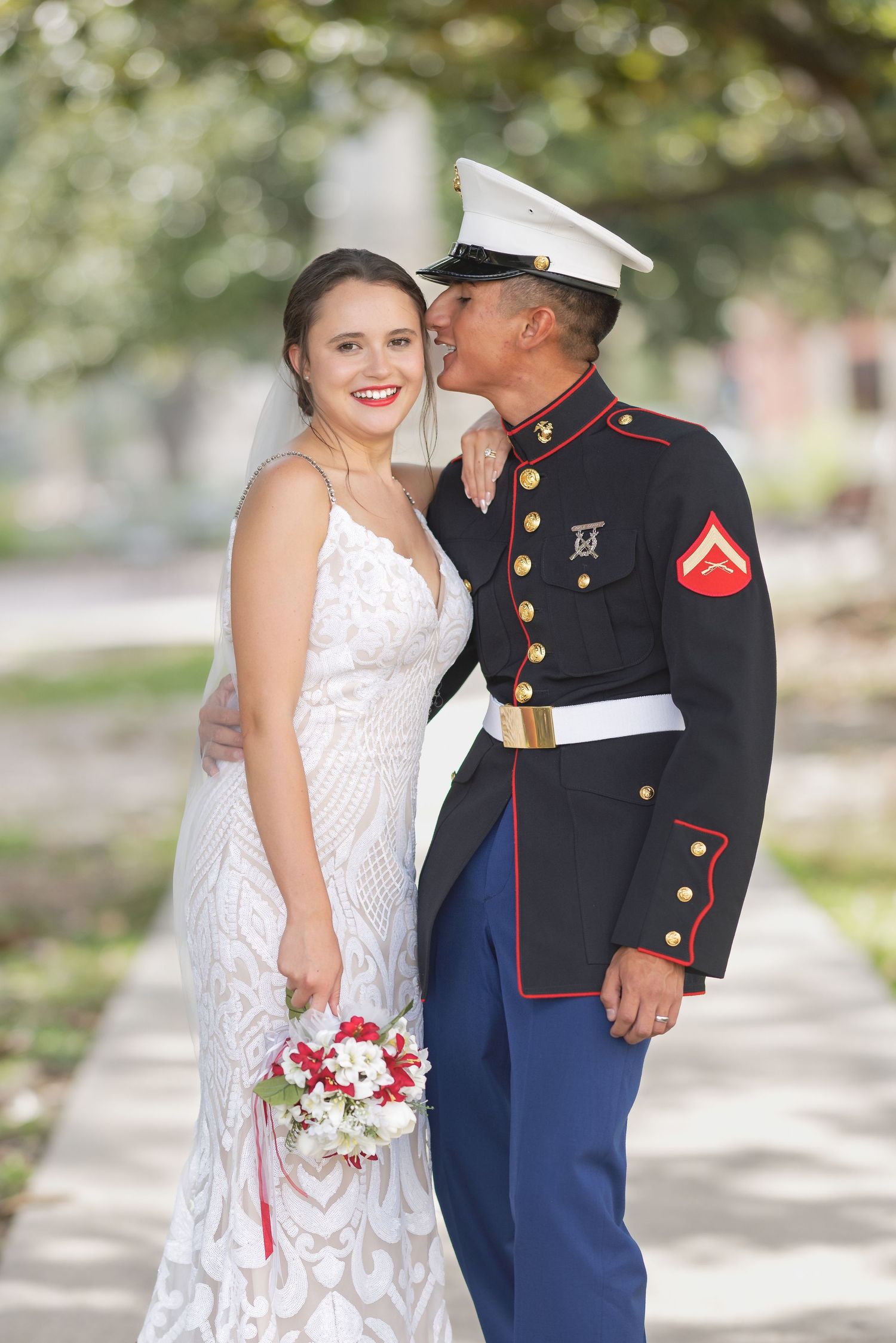 Marine in dress uniform kisses bride in white lace gown holding red and white bouquet outdoors.