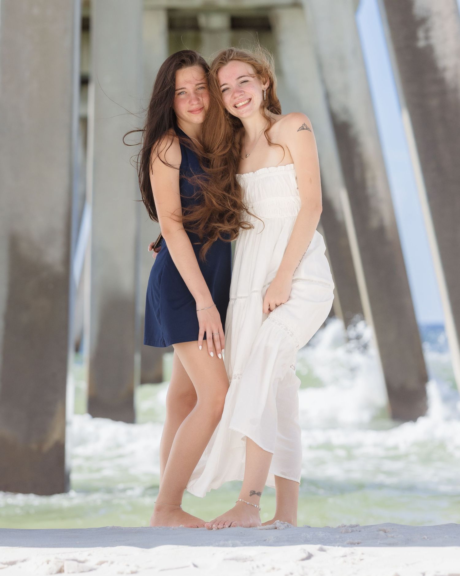 Two young women in navy and white dresses pose smiling on sandy beach under a pier.