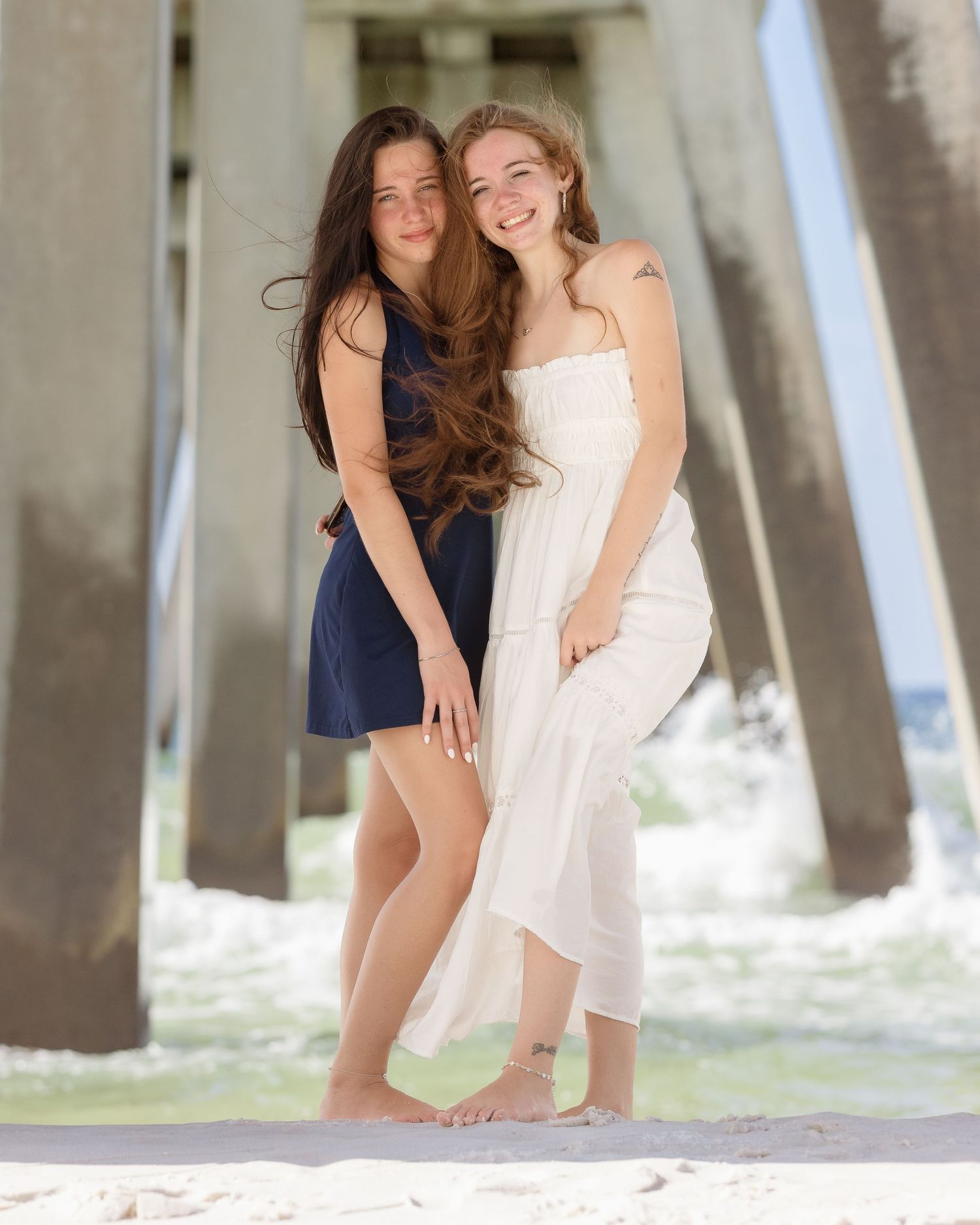 Two smiling young women in navy and white dresses posing together under a beach pier.