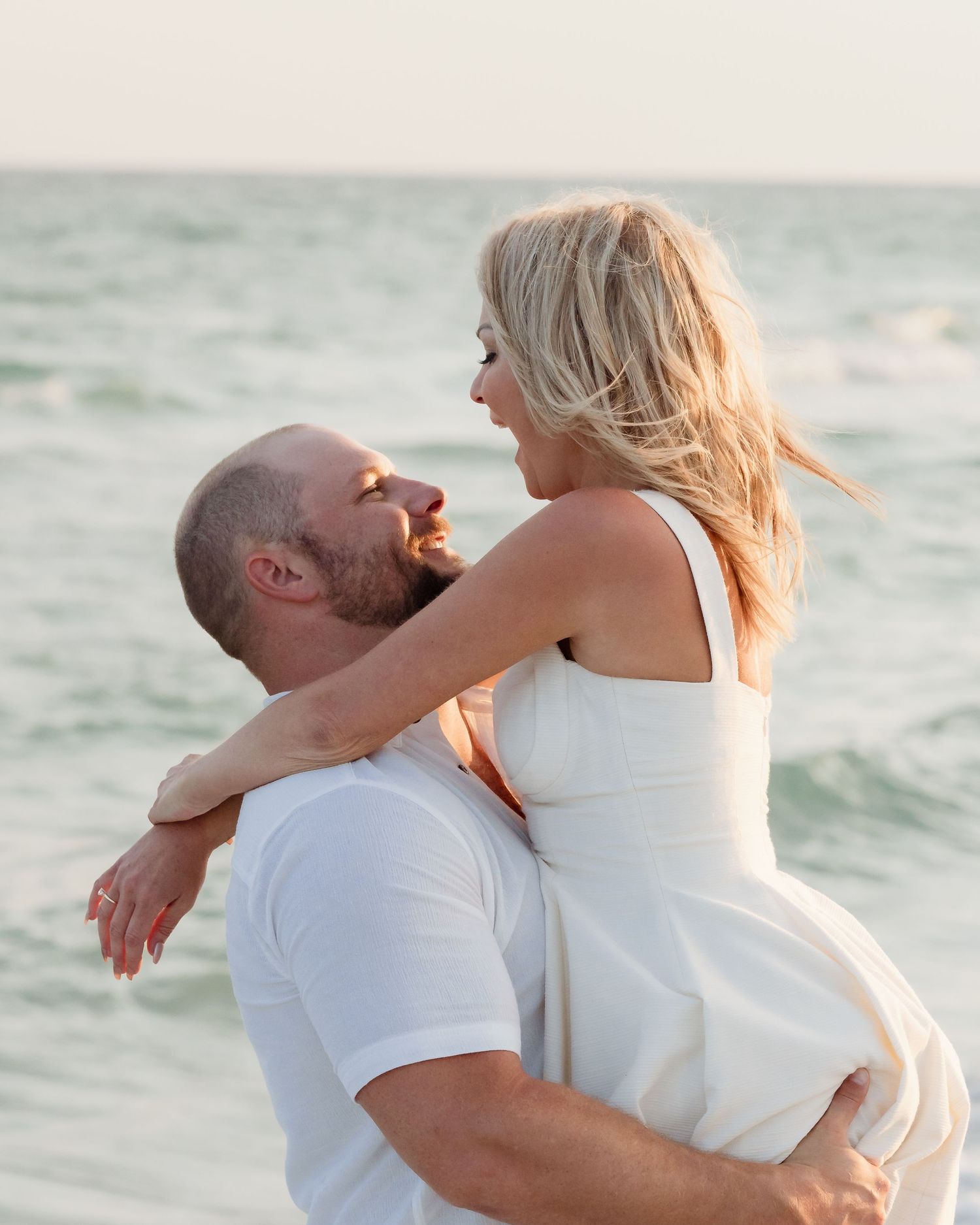 Man in white shirt lifting smiling blonde woman in white dress on ocean beach.
