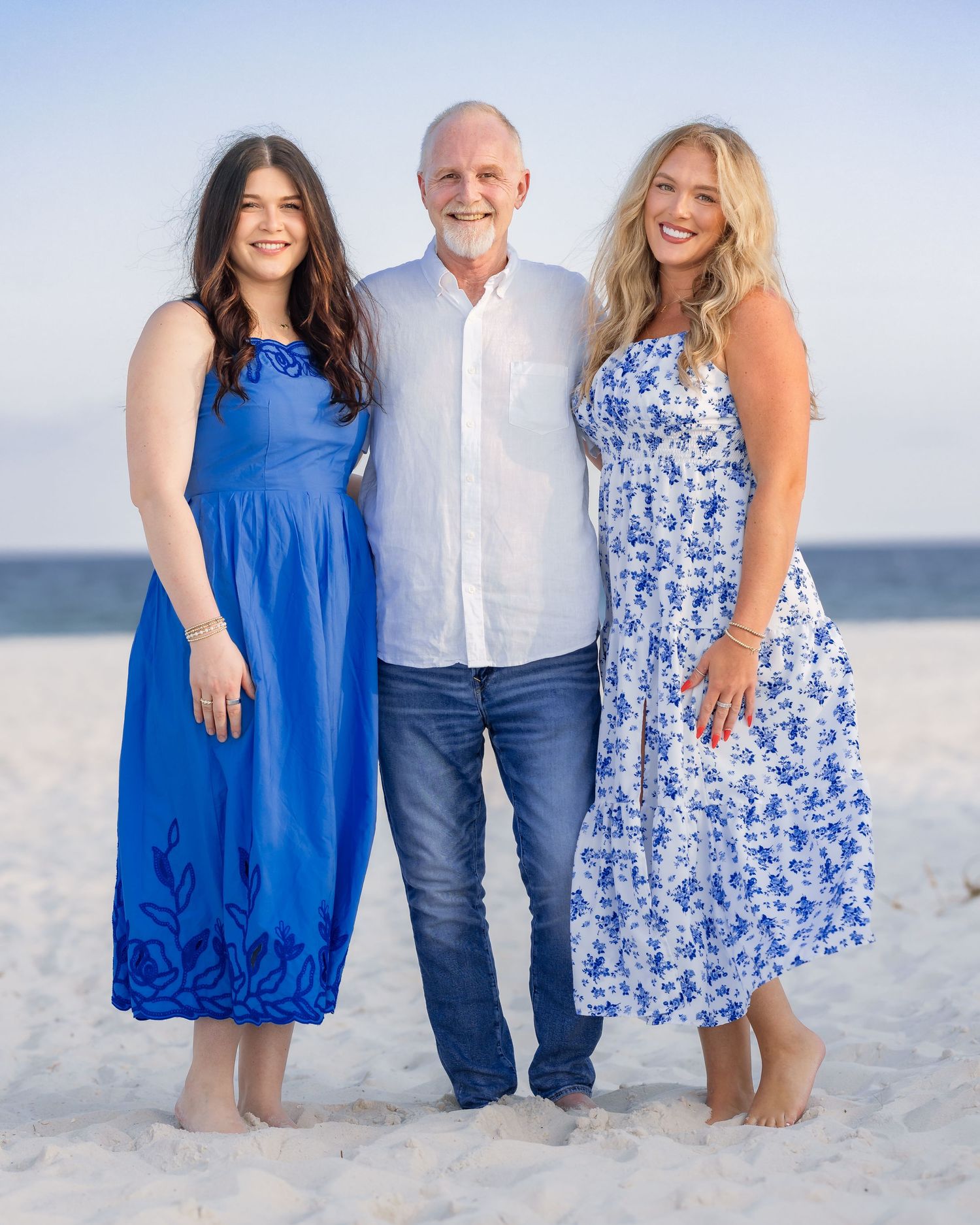 Family of three posing together on a beach, wearing coordinating blue and white outfits at sunset.