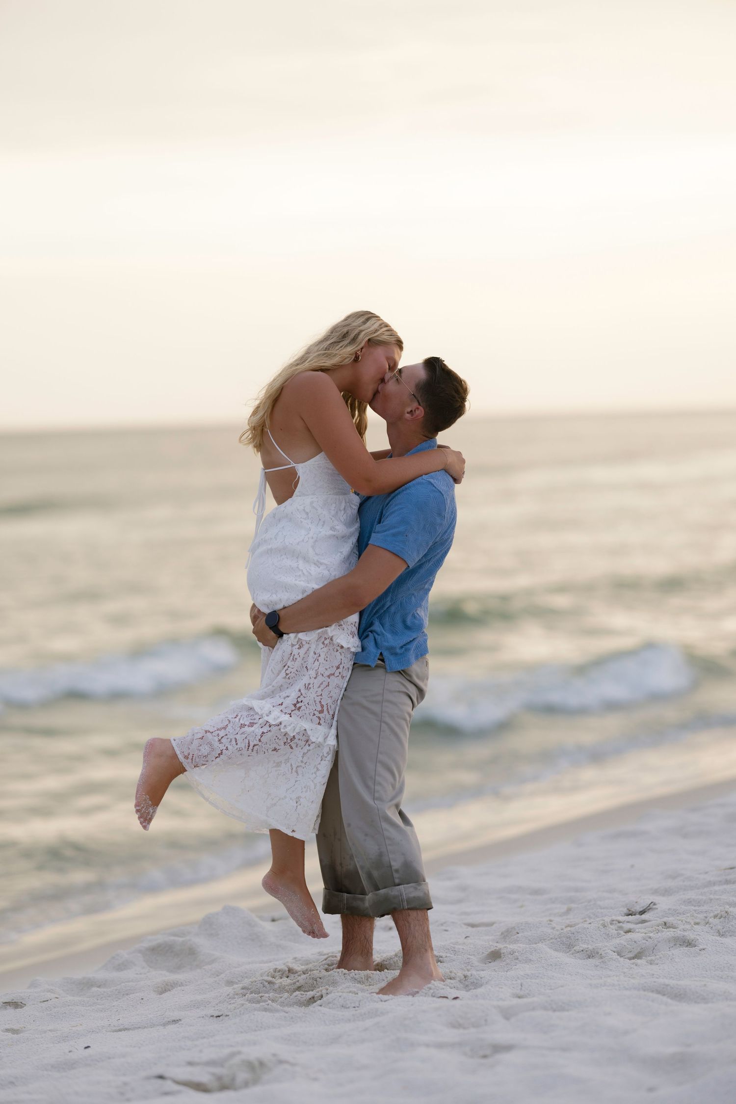 Couple embracing and kissing on a sandy beach at sunset, man lifting woman in white dress near ocean waves.