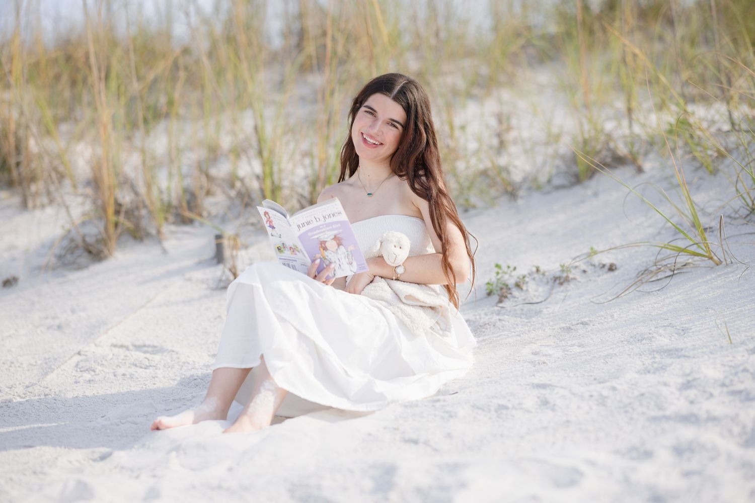 Young woman in white dress sitting on sandy beach dunes, smiling while holding a book on a sunny day.