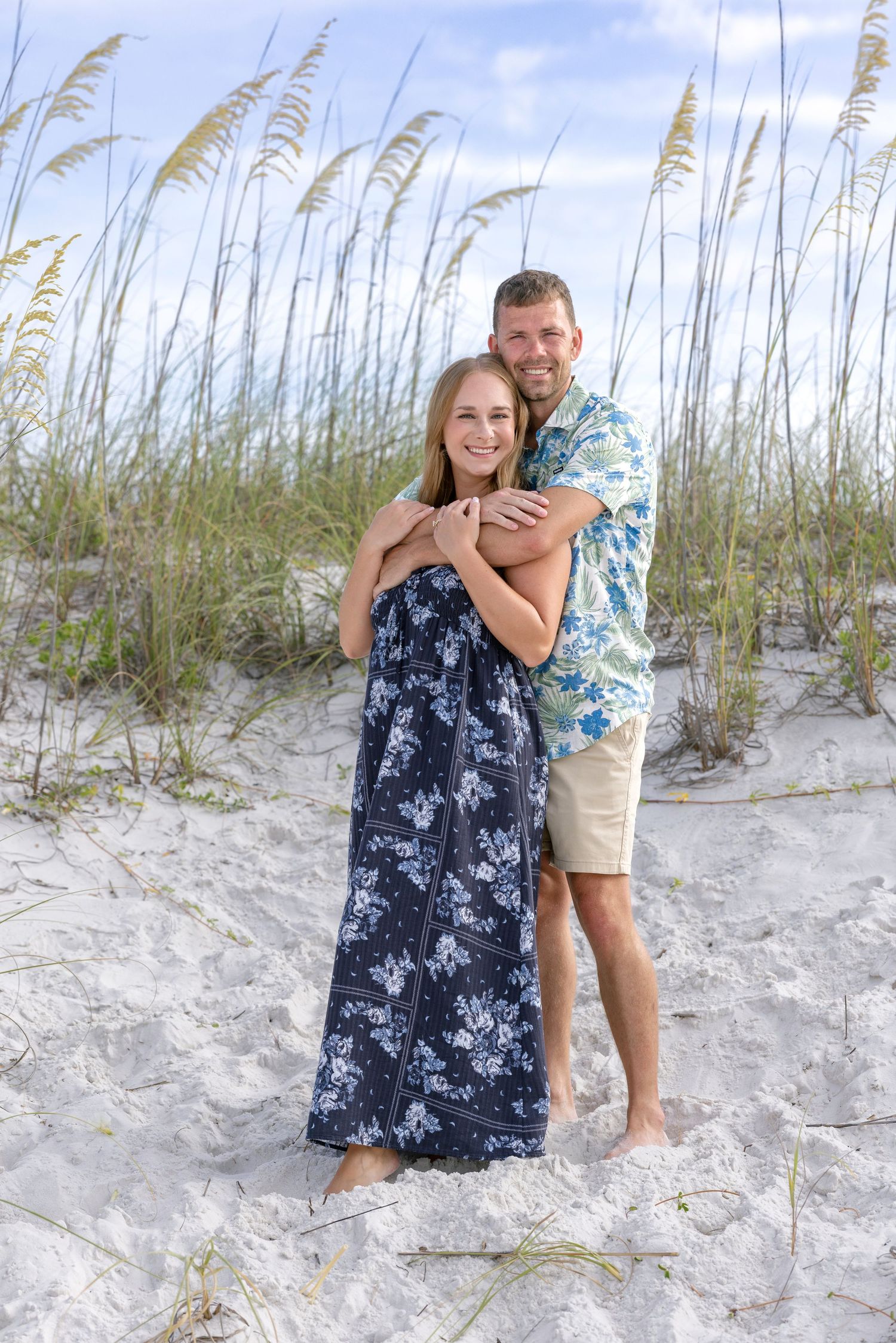Couple embracing on white sand beach dunes with sea oats, woman in navy floral dress, man in tropical shirt and shorts.