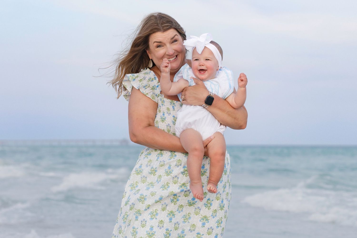 Smiling grandmother holding happy baby girl in white outfit on a breezy beach.