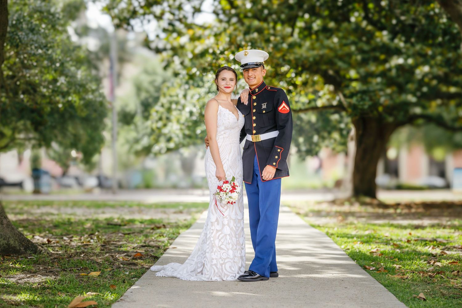 A bride in a white lace gown and a Marine in dress blues pose on a tree-lined path on their wedding day.