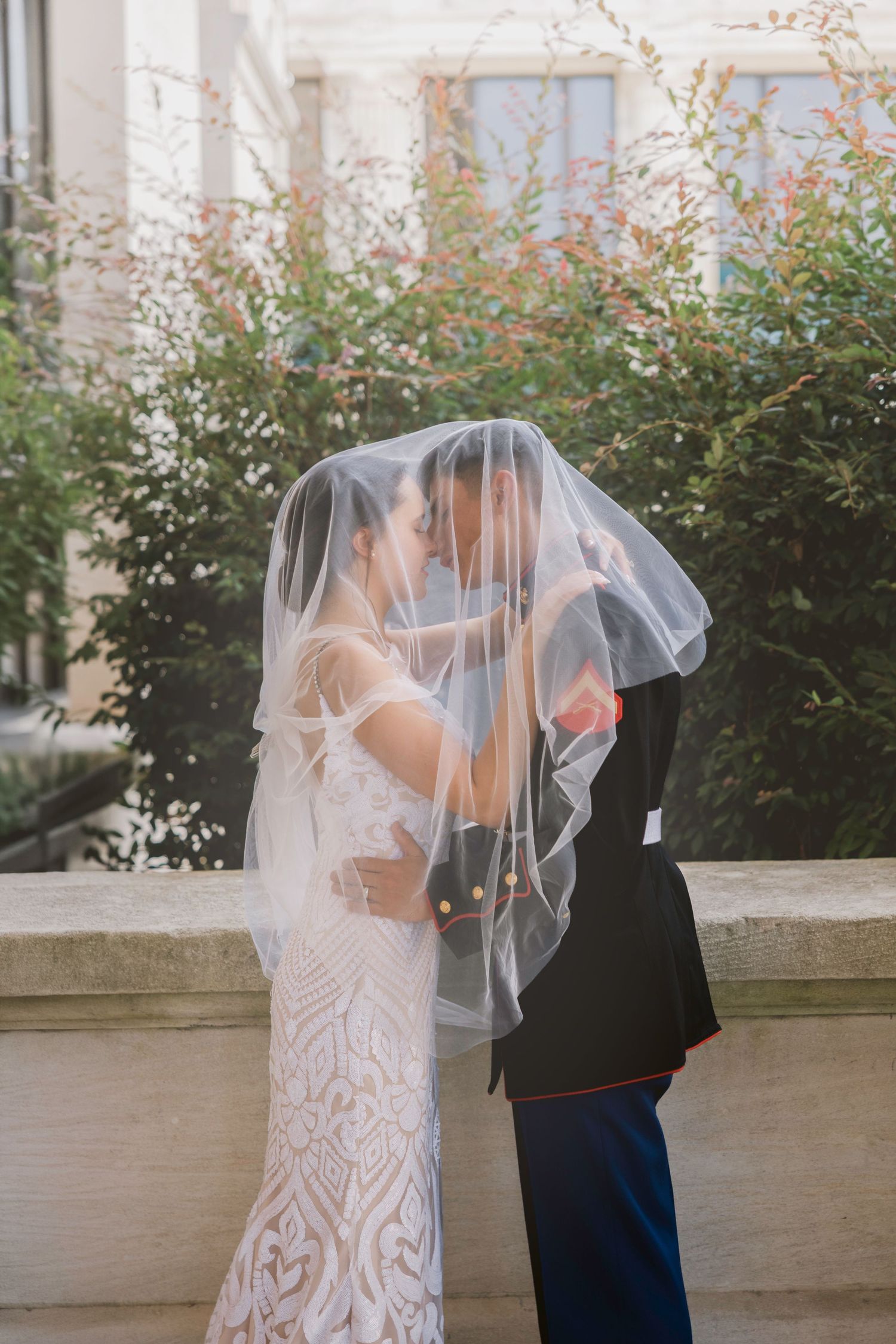 Bride and groom share romantic kiss under wedding veil outside historic building with lush greenery.