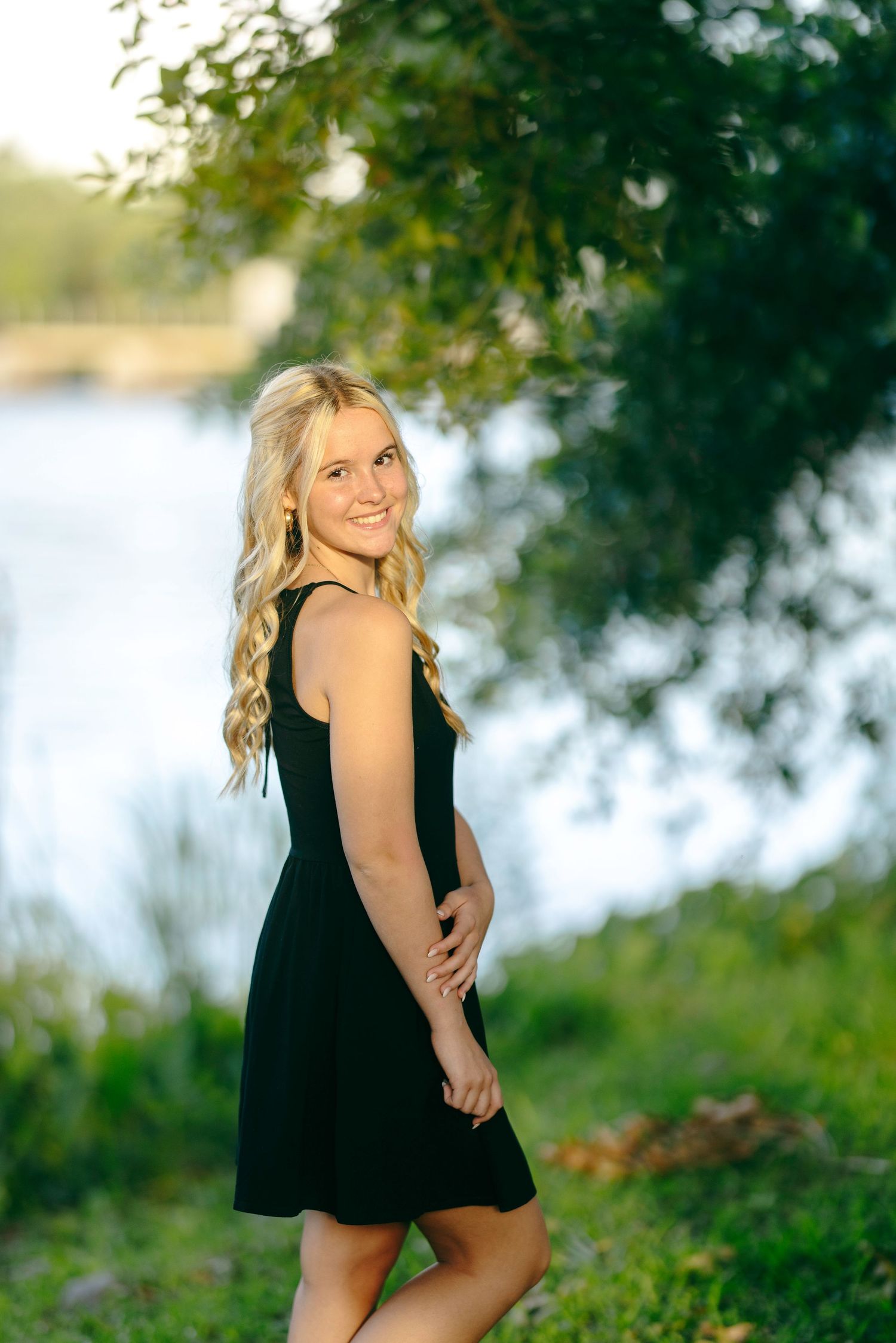 Outdoor summer photoshoot of woman in black dress posing by water with trees in background at sunset.