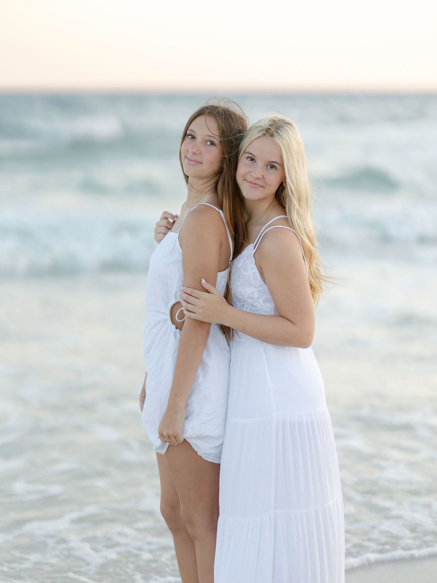 Two people in white dresses pose back-to-back on a beach at sunset with ocean waves in the background.