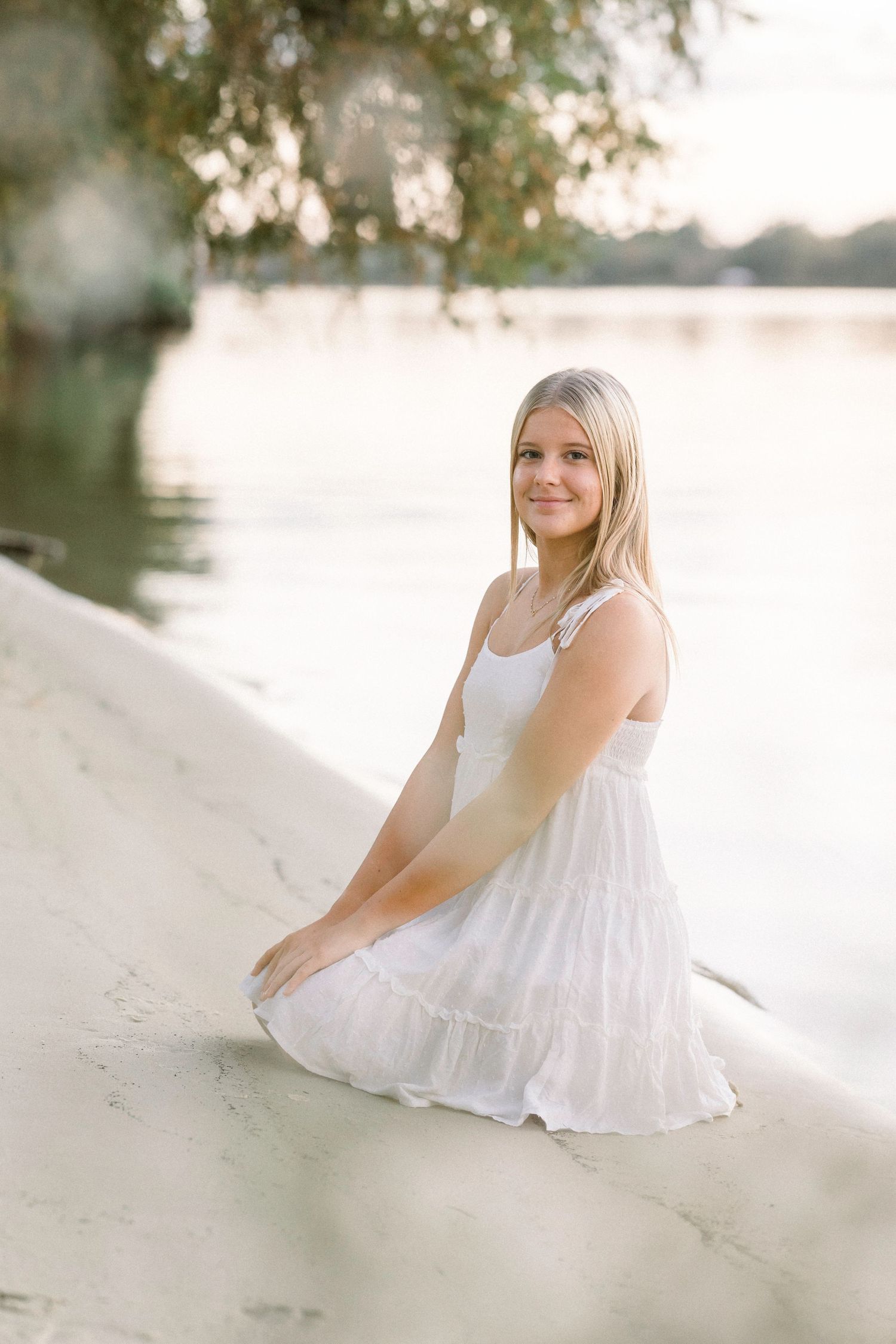 Blonde girl in white sundress sitting on sandy shore by calm lake in golden hour light.