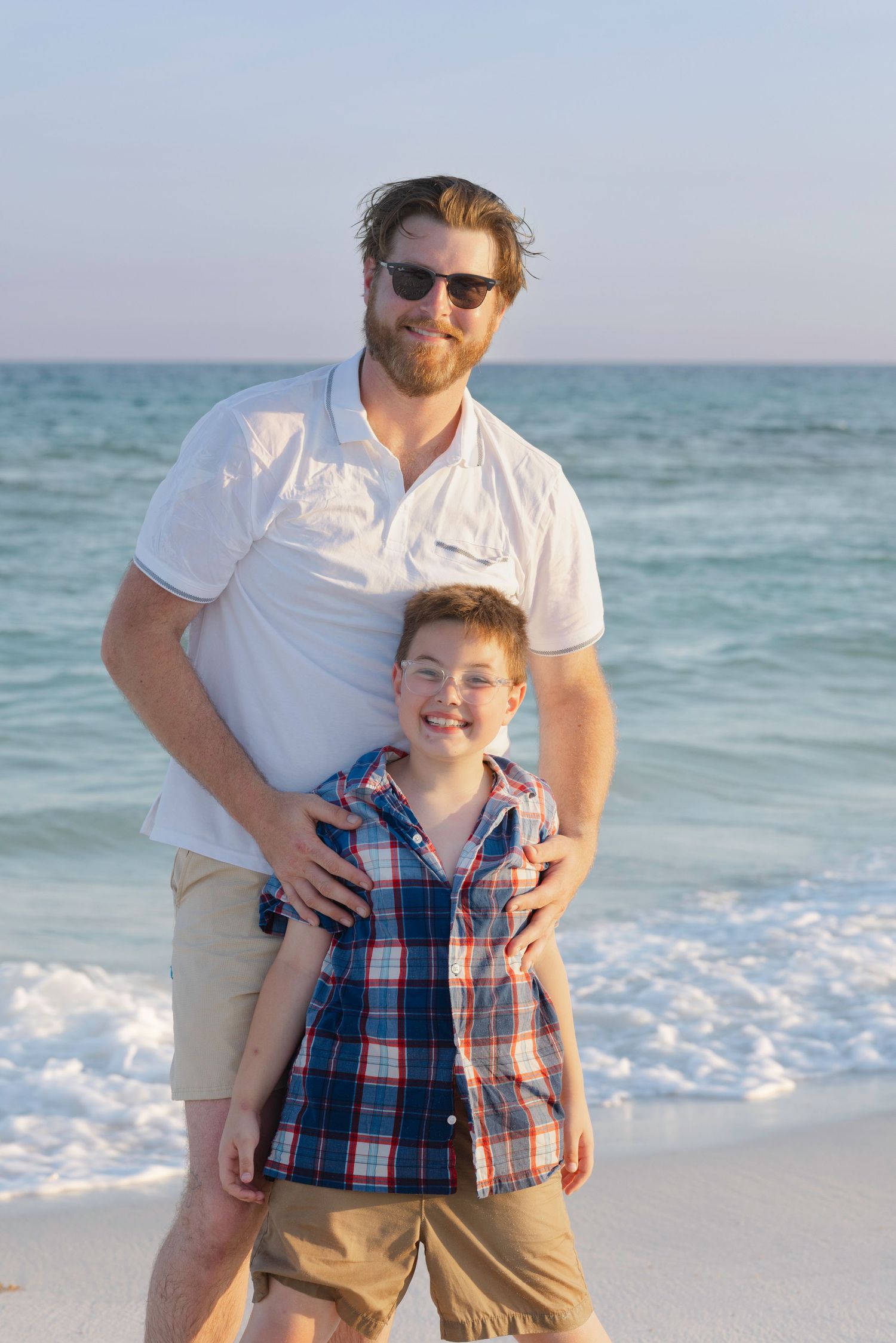 A father and son smile together on a sunny beach, with ocean waves in the background, both wearing casual summer outfits.