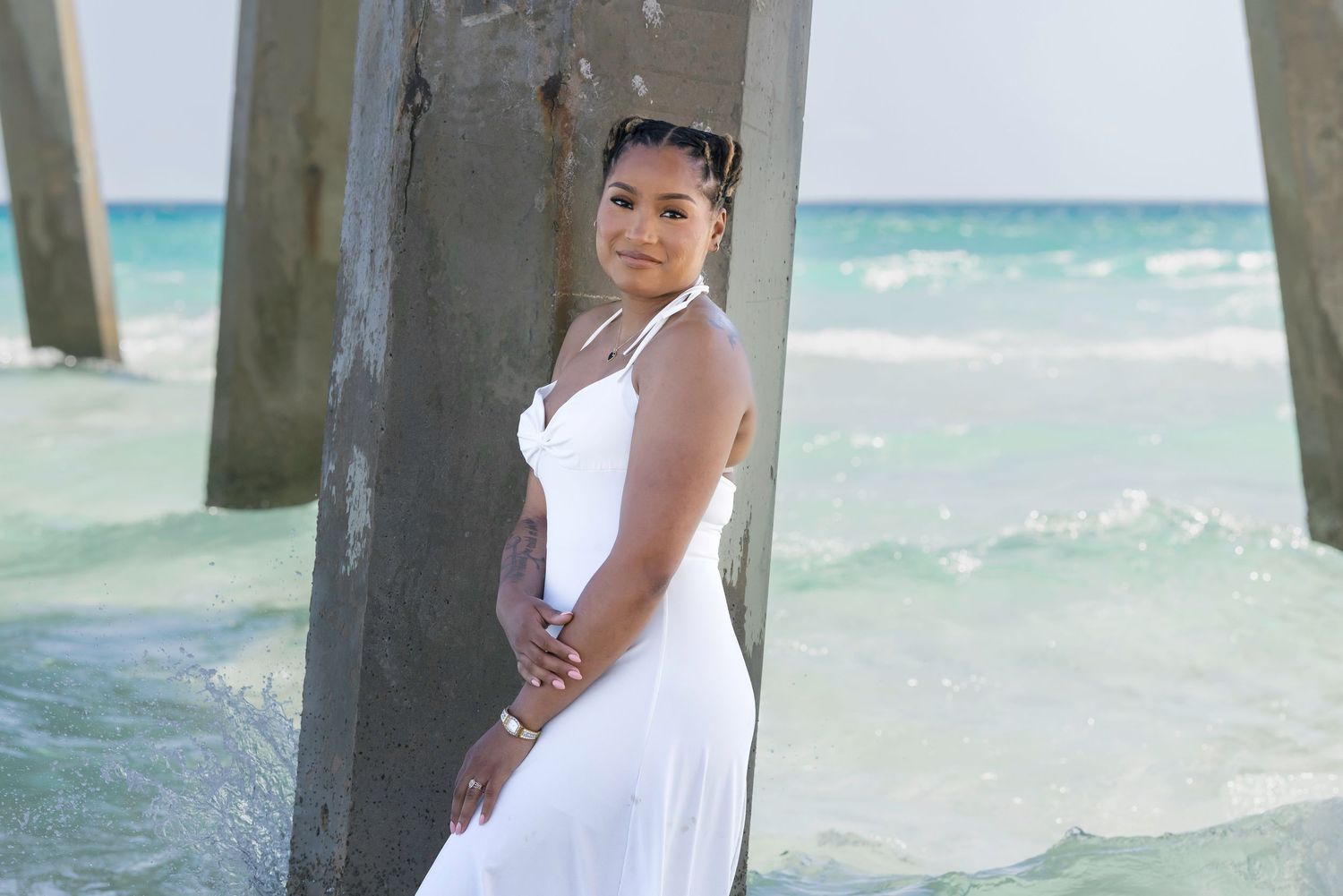 Woman in white dress posing by pier pillar on tropical beach with turquoise water.