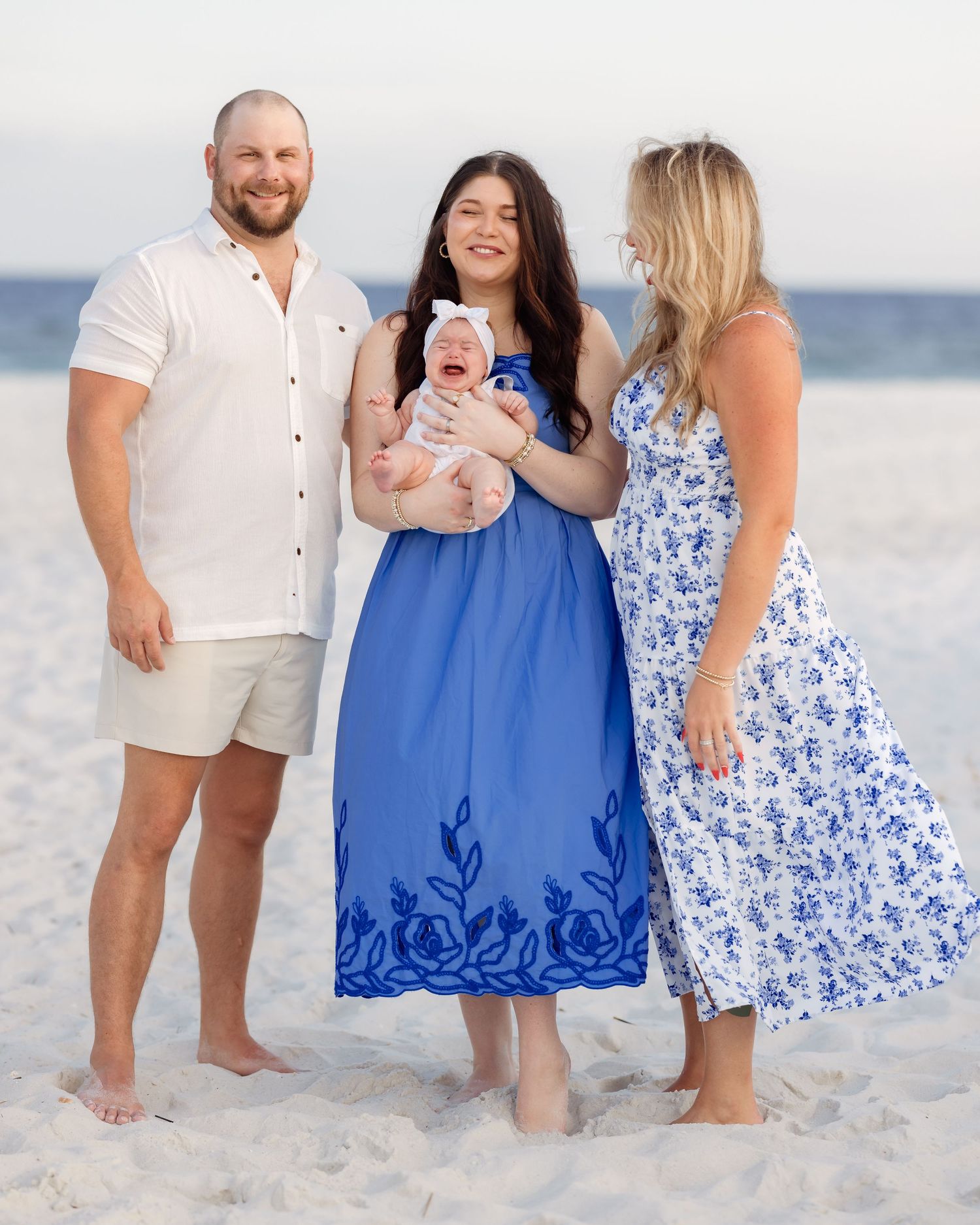 Family of three posing on a sandy beach, woman in blue floral dress holding newborn baby, man in white shirt and khaki shorts.