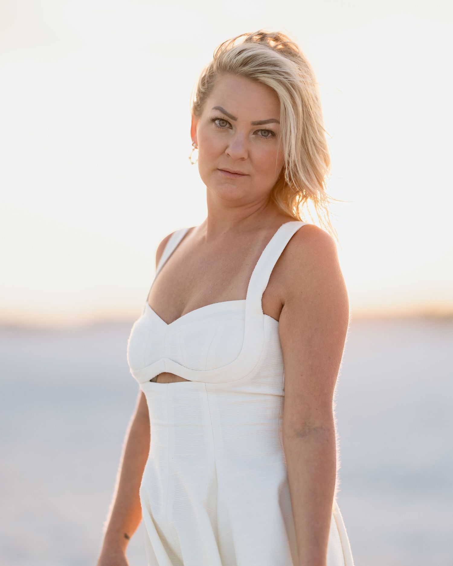 Blonde woman in white sundress poses at golden hour on a serene beach at sunset.