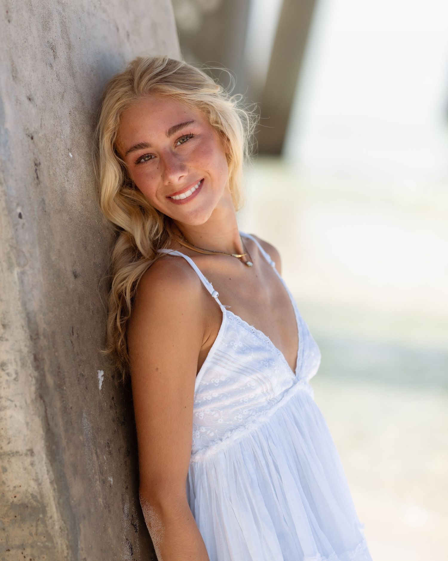 Smiling blonde woman in a white sundress leaning against a concrete pillar outdoors on a sunny day.