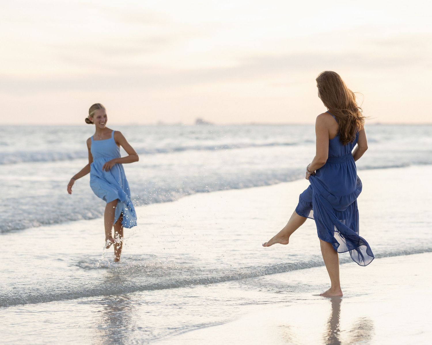 Two women in blue dresses running and playing along a serene beach shoreline at sunset.