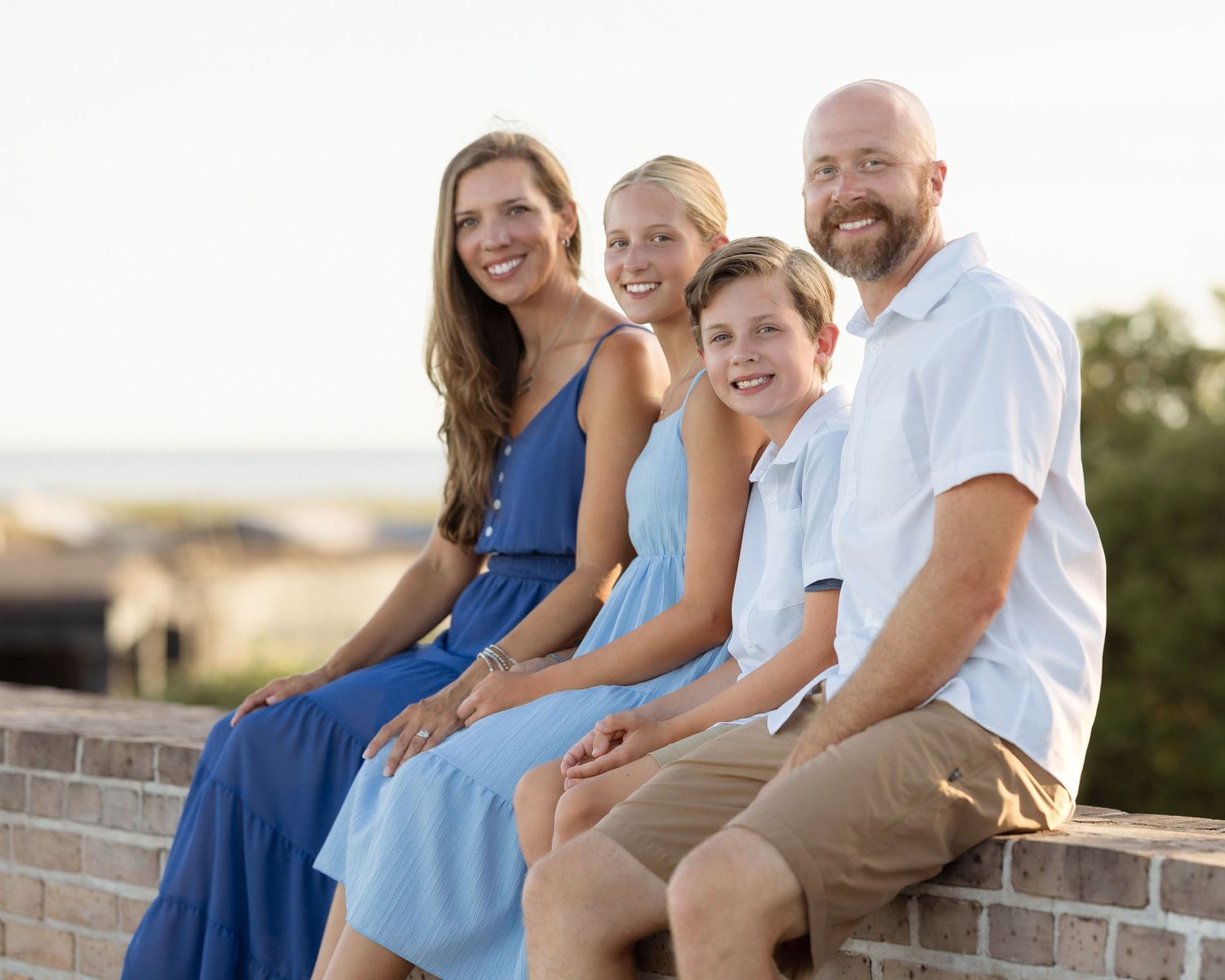 Happy family of four posing together on a brick wall with a scenic beach backdrop on a sunny day.