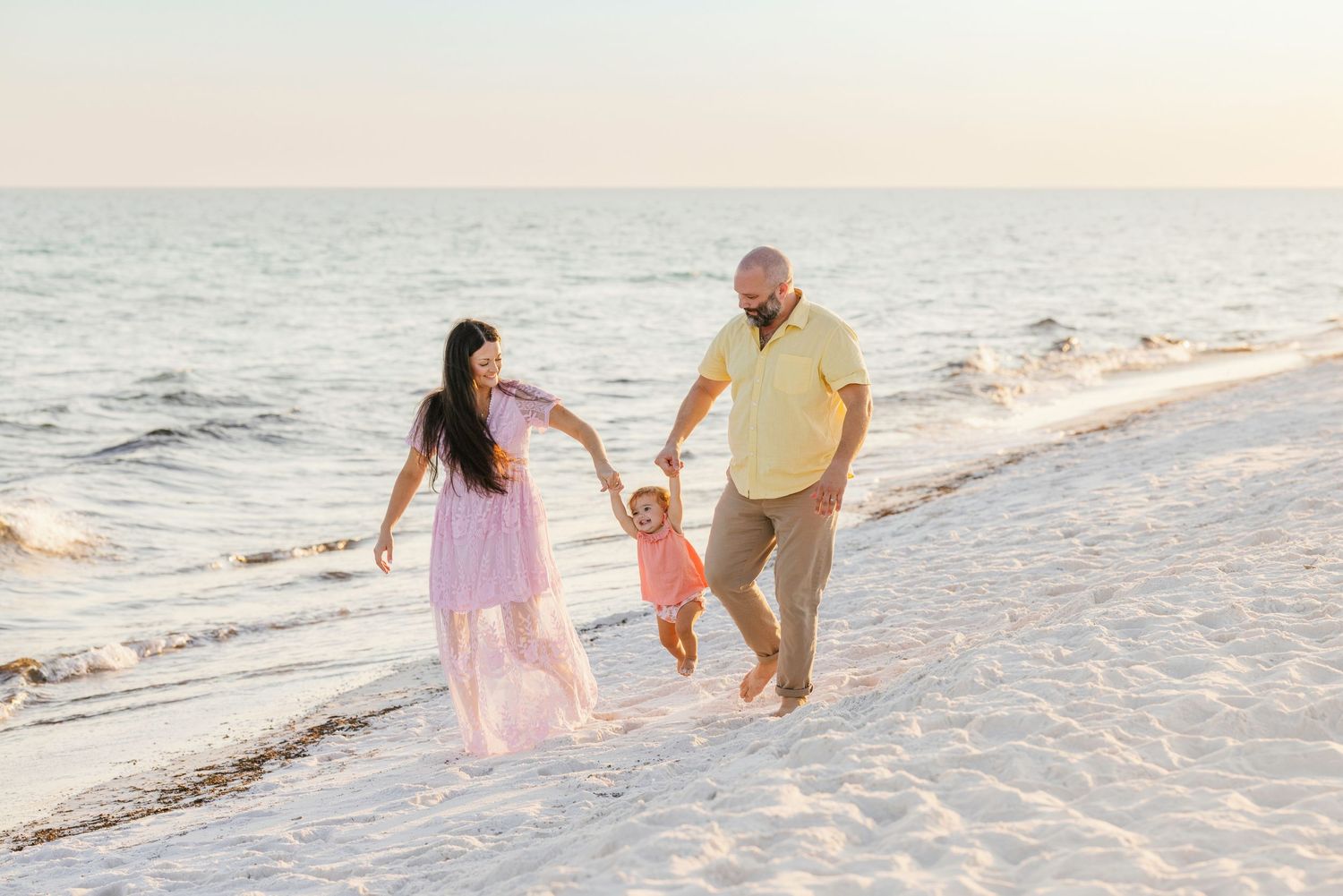 A family in casual attire walks along a serene beach at sunset, with waves gently lapping at the shoreline.