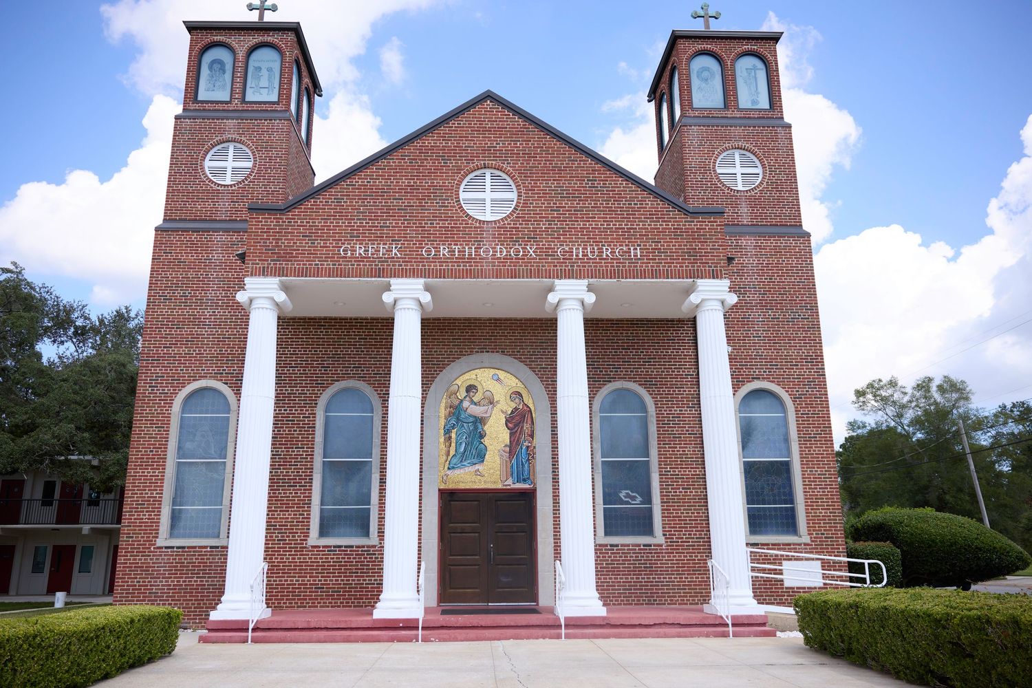 A row of red brick church buildings with white columns and steeples photographed against a bright blue sky with clouds.
