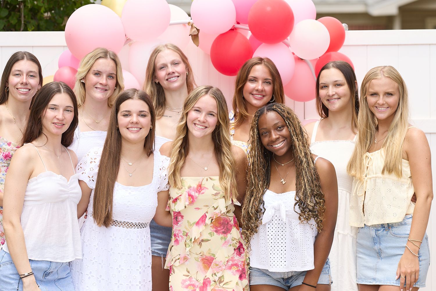 A group poses outdoors with pink and red balloons against a white fence on a sunny day.