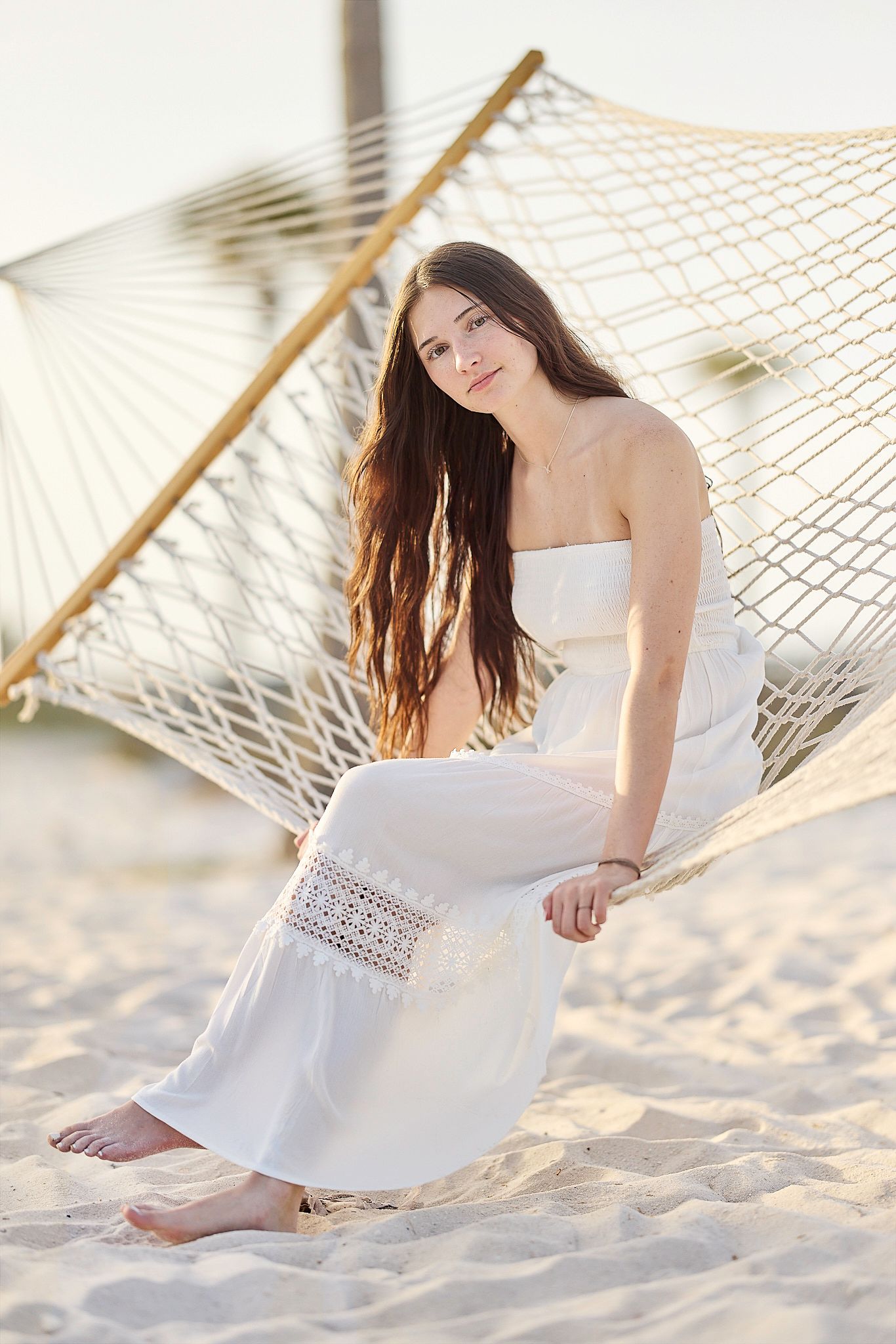 Casual beachside portrait with hammock and flowing white dress against sandy backdrop.