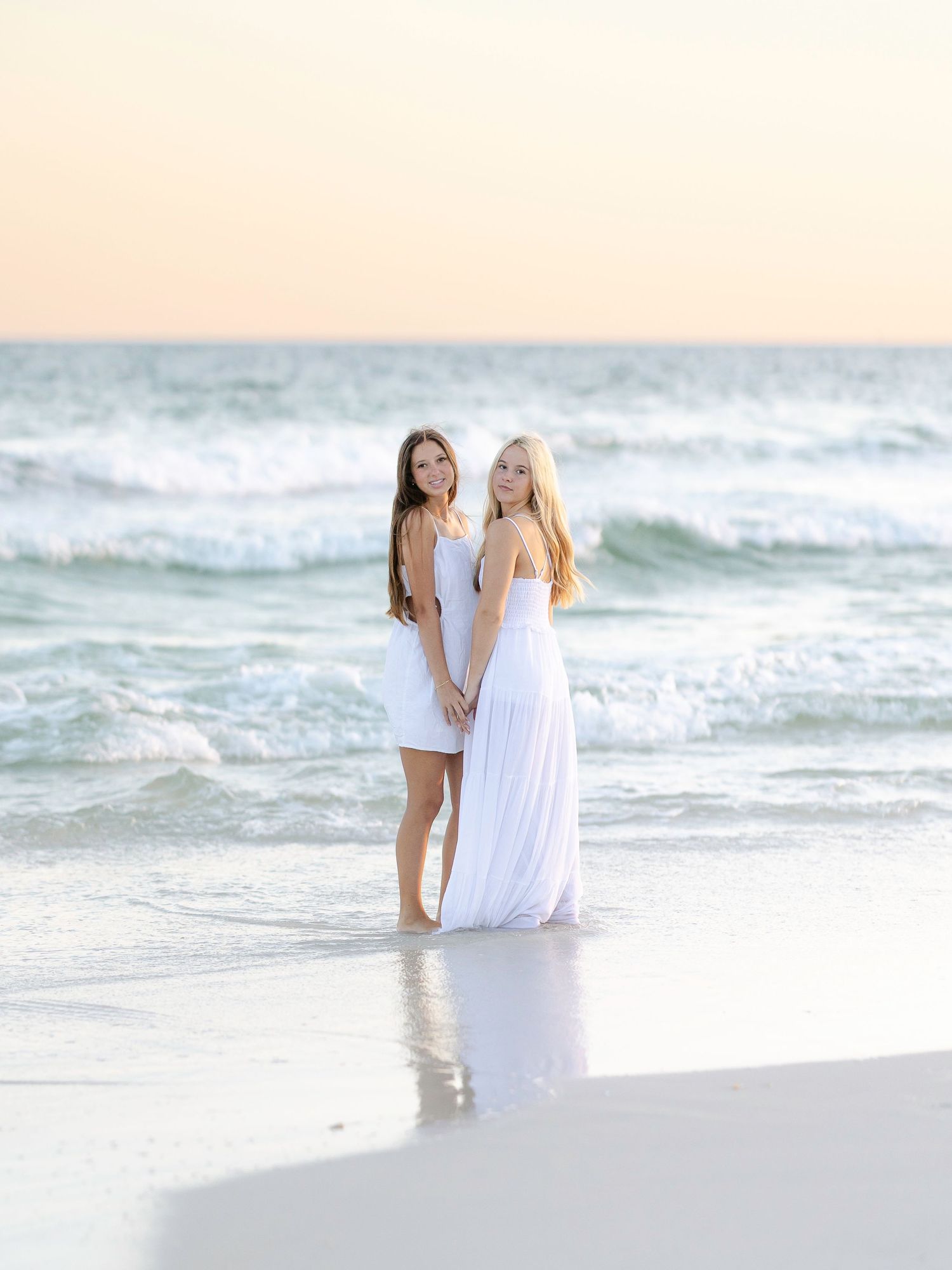 Two people in white dresses walking along shoreline at sunset.