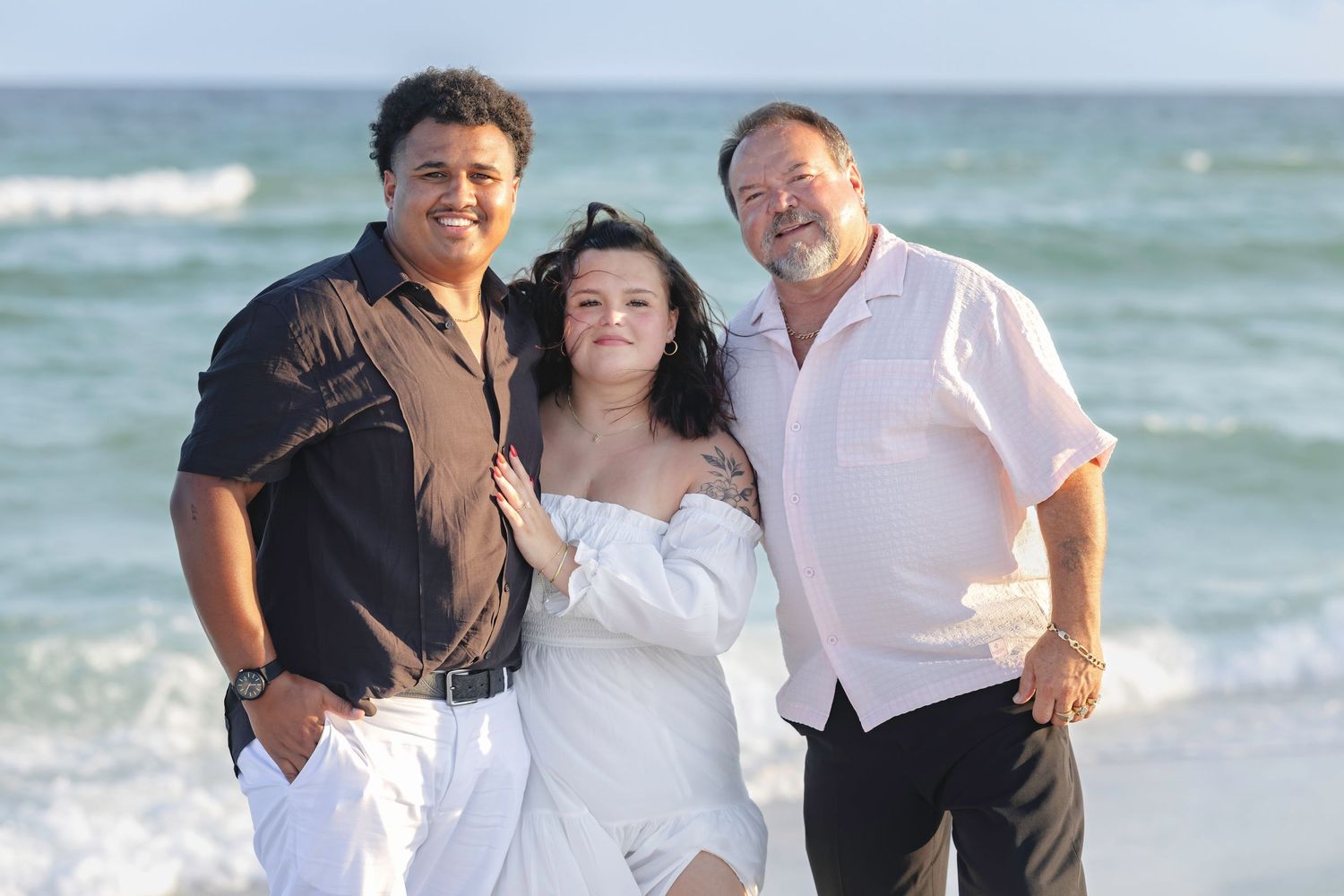 Three people smiling together on a sunny beach, with ocean waves in the background, dressed in white and black attire.