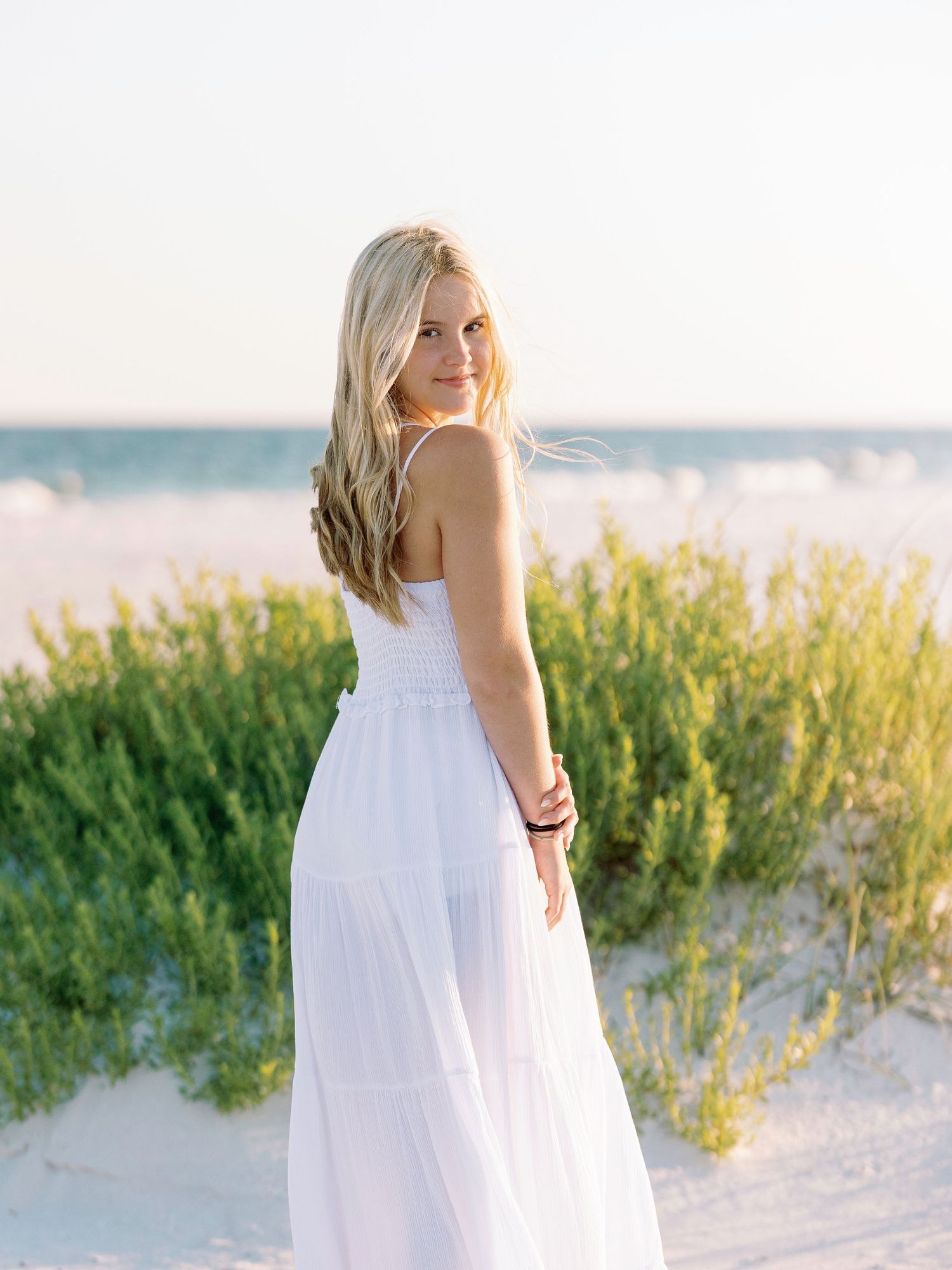Young blonde woman in white sundress posing on beach sand dunes with ocean view behind her.