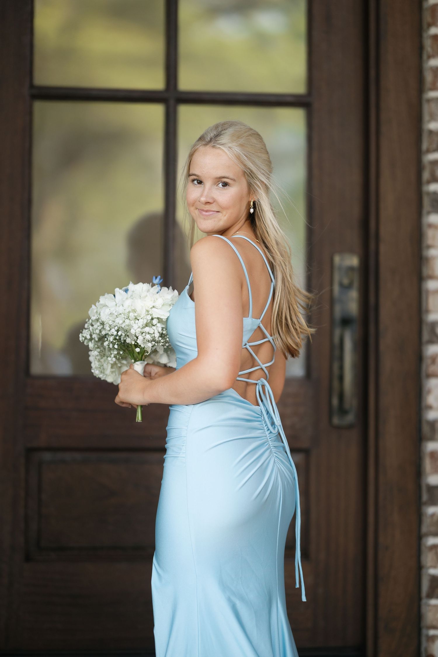 Blonde woman in a light blue lace-up back mermaid dress holding white flowers by a wooden door.