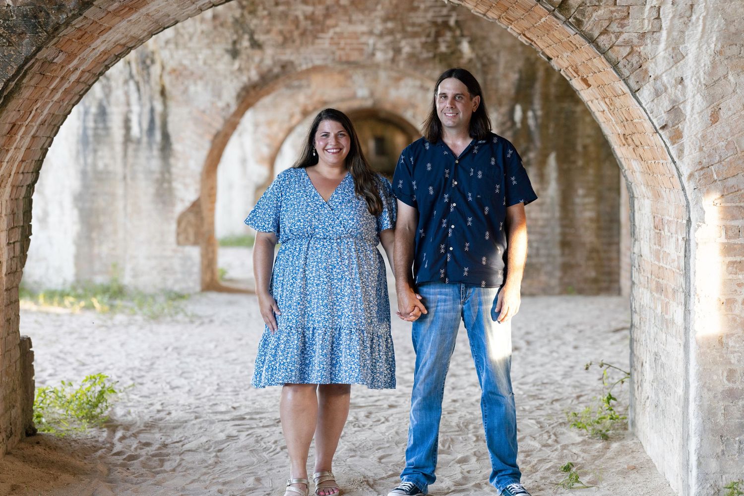 Couple holding hands under brick arches at historic fort, woman in blue floral dress and man in navy shirt with jeans.