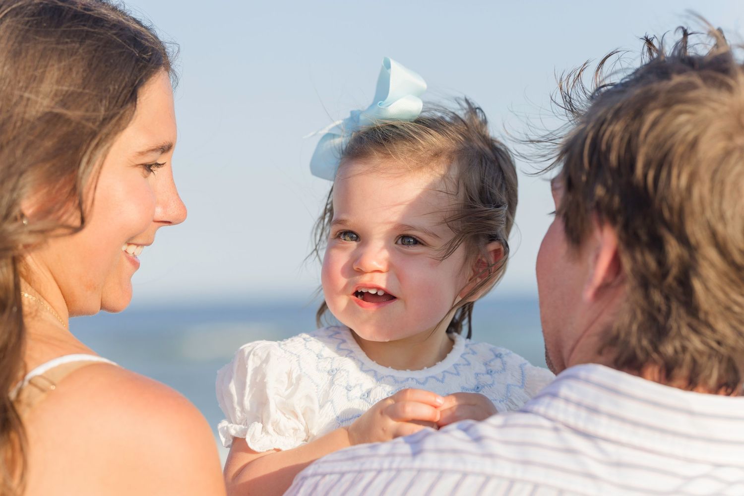 Happy parents smiling at their toddler daughter with a blue bow on a sunny beach.