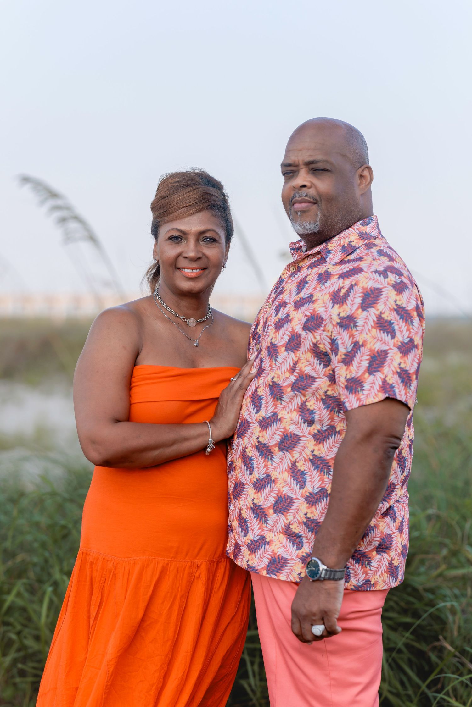A couple poses outdoors near beach grass, woman in orange dress, man in pink floral shirt and pink pants.