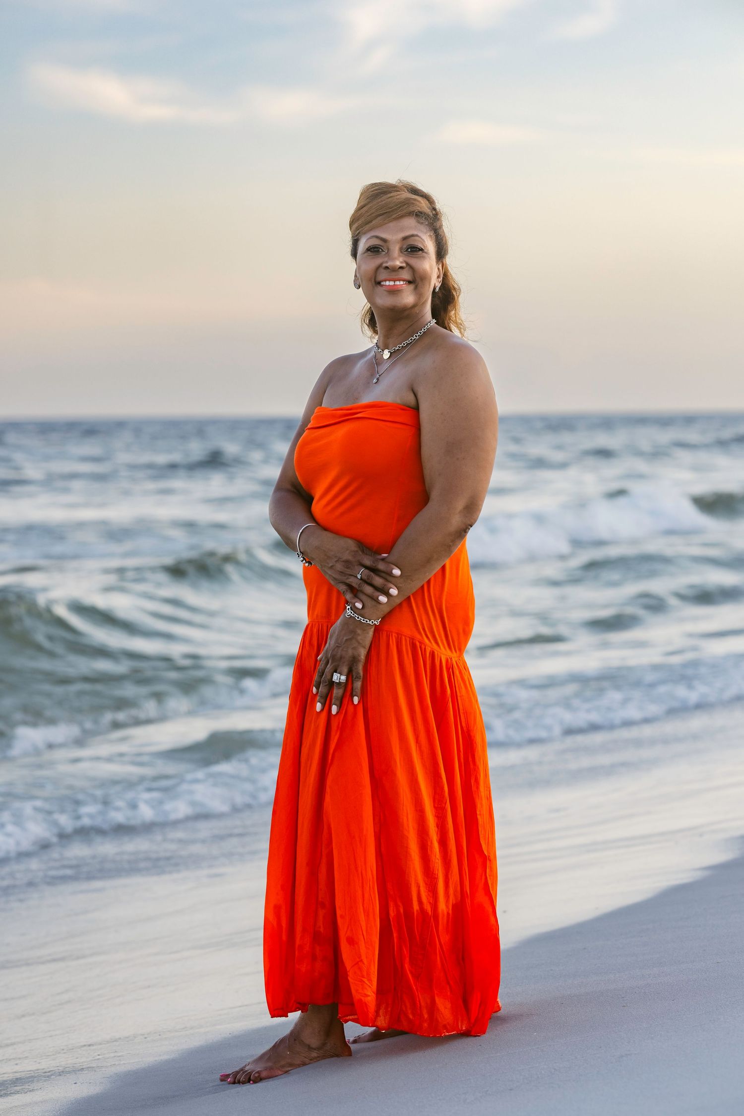 A smiling woman in an orange strapless maxi dress stands barefoot on a sandy beach with ocean waves behind her.