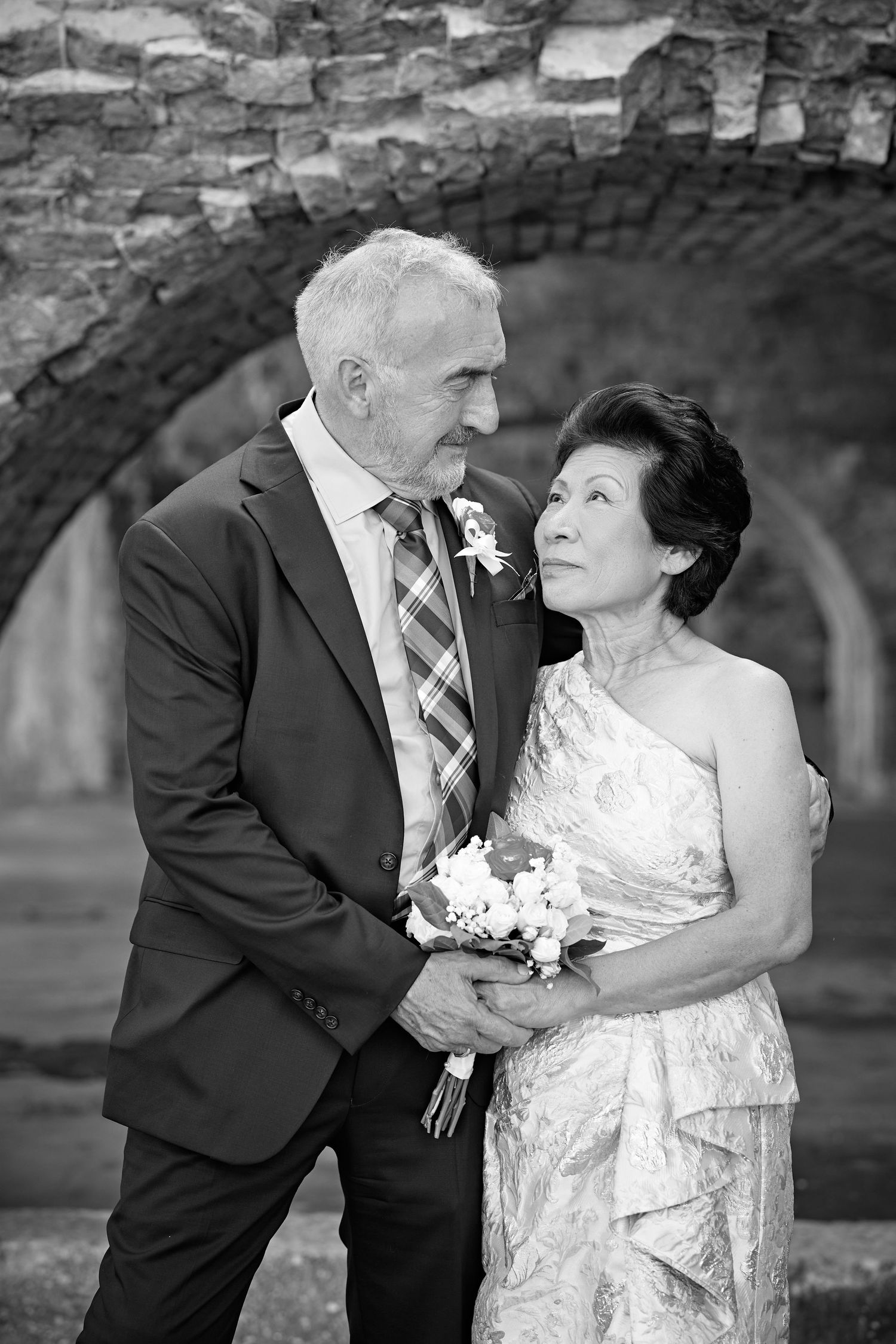 Black and white photo of an elegant older couple in formal attire holding a bouquet under stone arches.