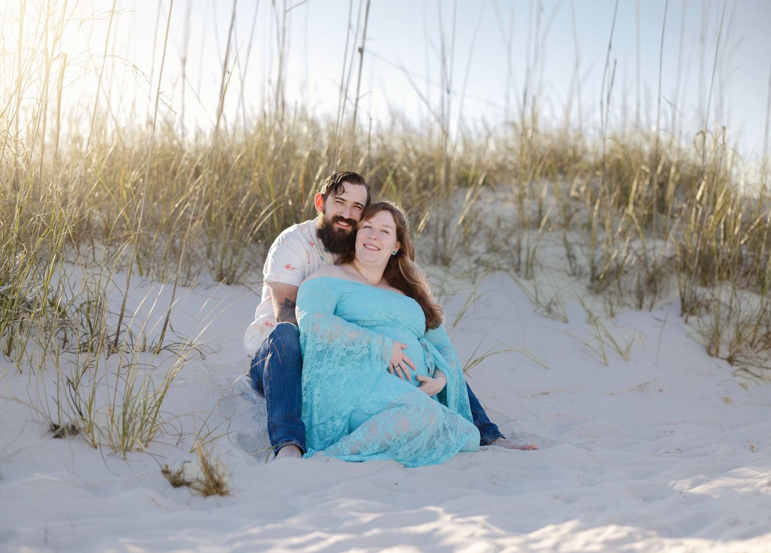 Pregnant couple sitting on white sand dunes with tall grass, woman in teal maternity gown, man embracing her from behind.