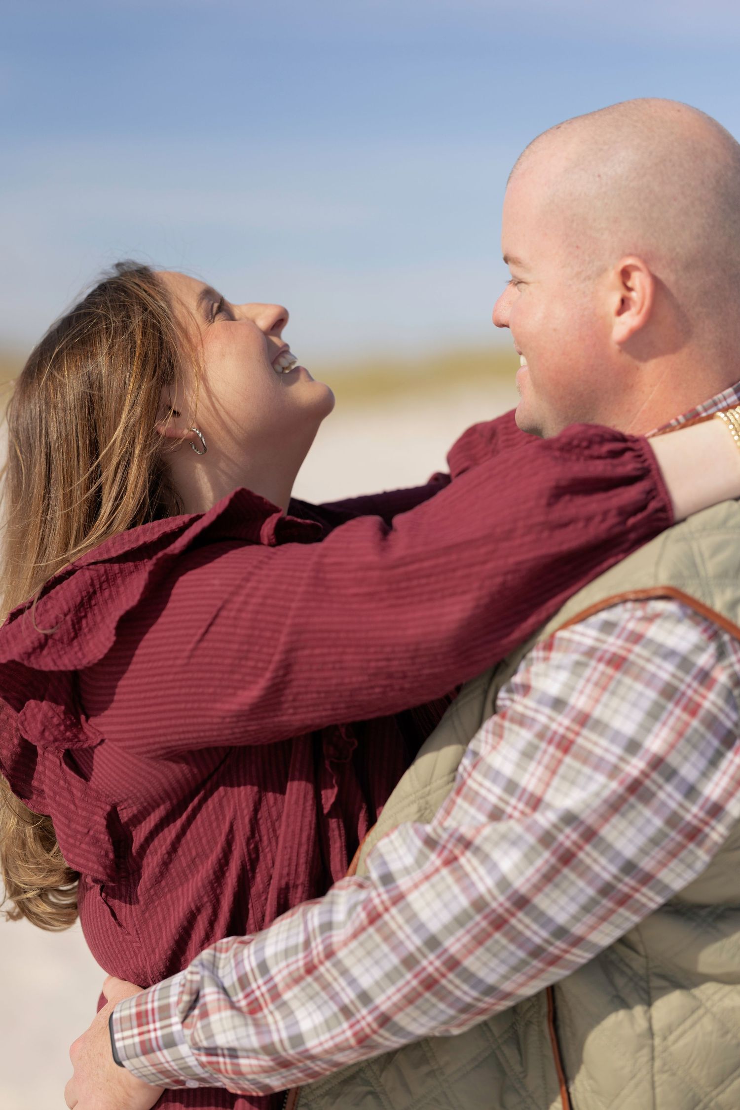 Joyful couple laughing and embracing on a bright sandy beach, woman in burgundy dress and man in green vest.
