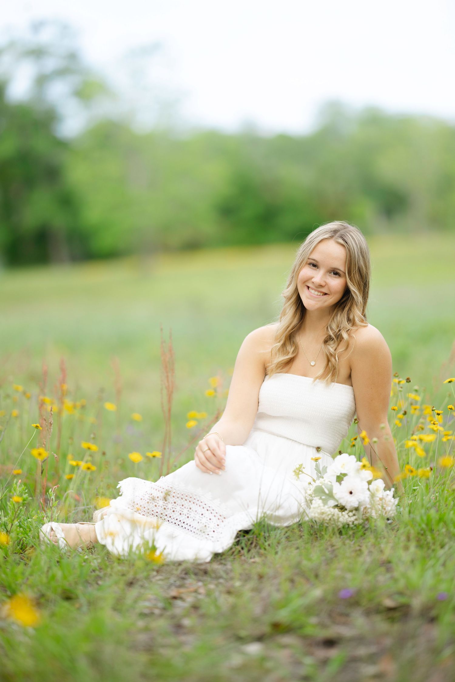 Blonde woman in white strapless dress sitting in a wildflower meadow with white flowers nearby.