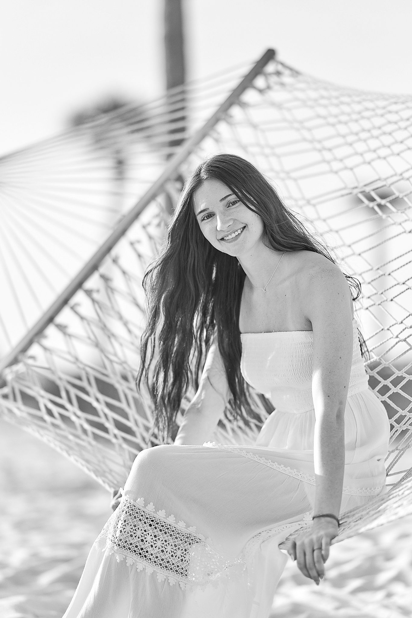 Black and white photo of smiling young woman in white dress sitting in rope hammock on beach.