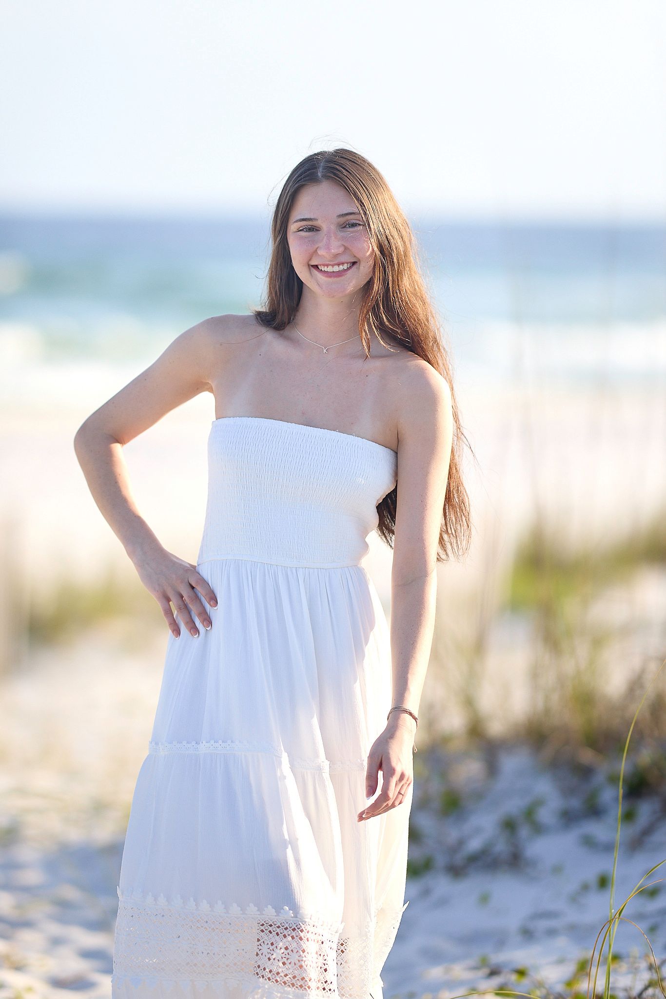 Young woman in white strapless maxi dress smiling on a sunny beach with ocean waves in the background.