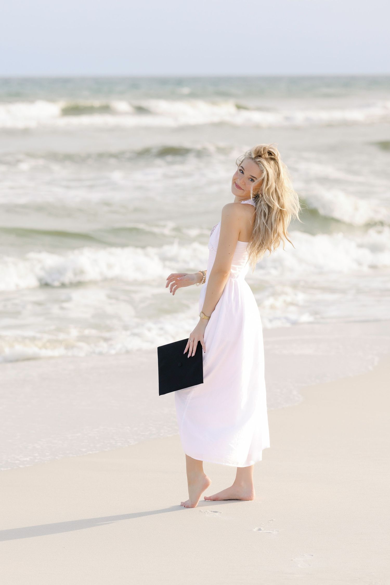 Blonde woman in white dress holding graduation cap on sandy beach with ocean waves.