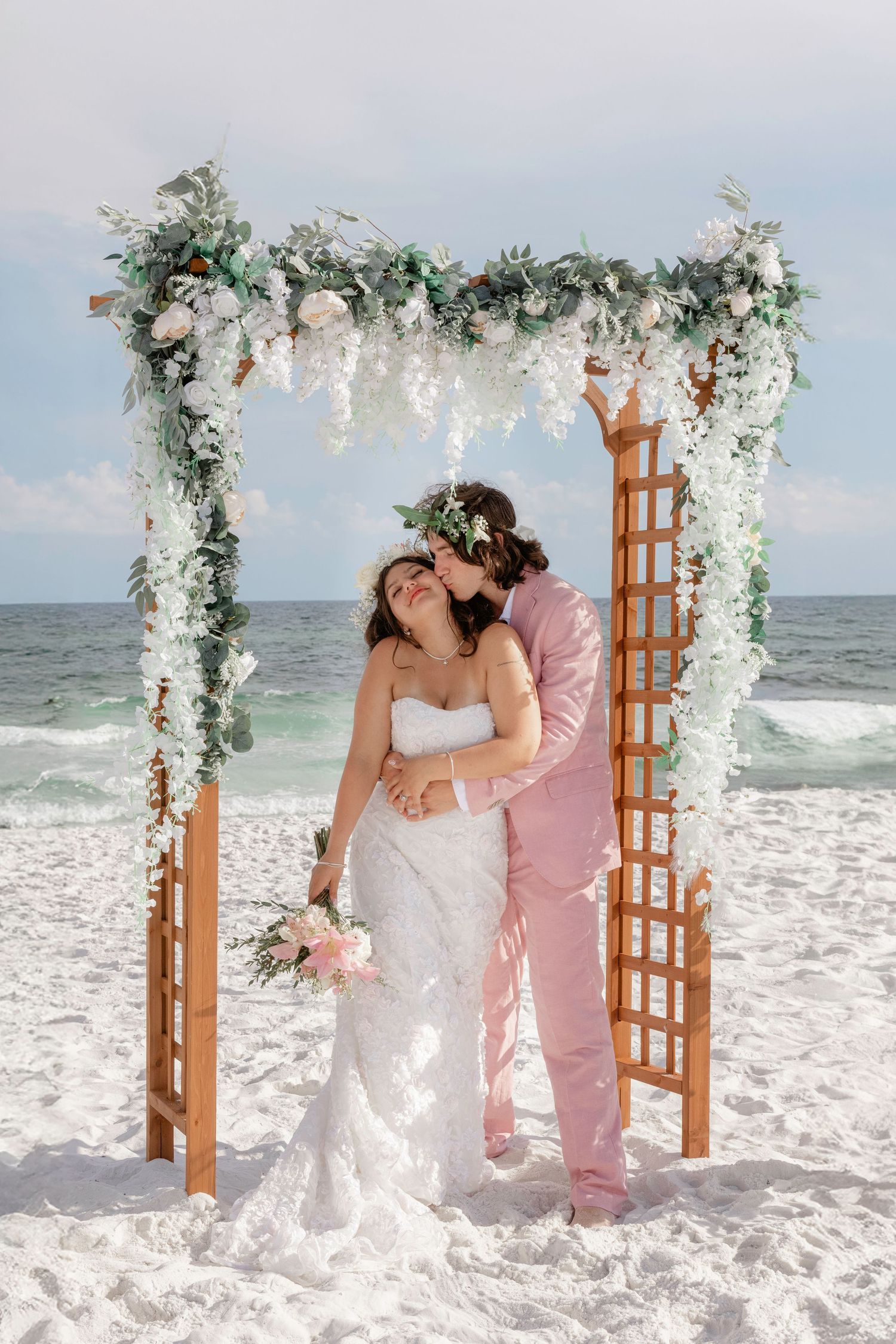 Couple sharing a kiss under a floral arch on a white sand beach, bride in white gown holding pink bouquet.
