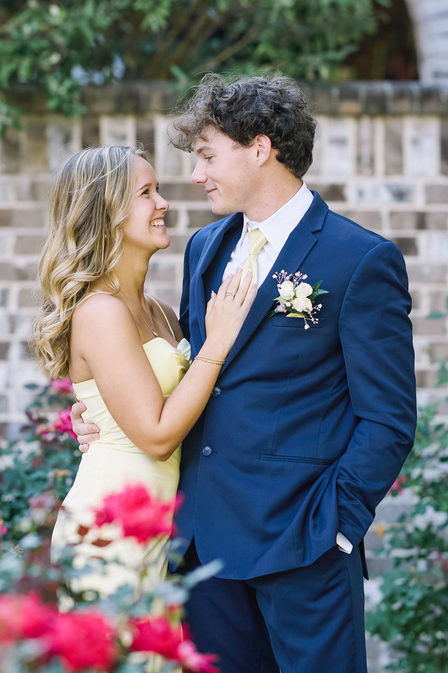 A couple in formal attire share a romantic moment in a garden with blooming red flowers in the background.