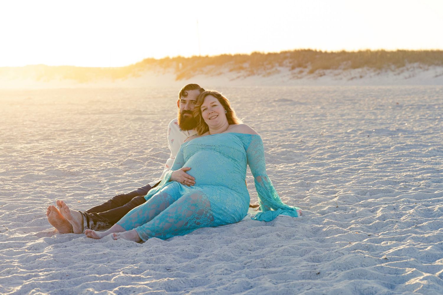Couple sitting on sandy beach at golden sunset, pregnant woman in teal dress cradling baby bump.