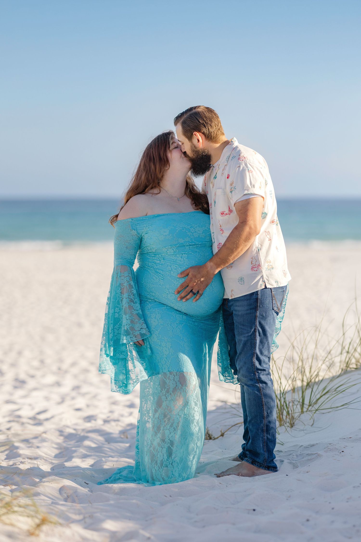 Bearded man kissing pregnant woman in turquoise lace maternity dress on white sand beach.