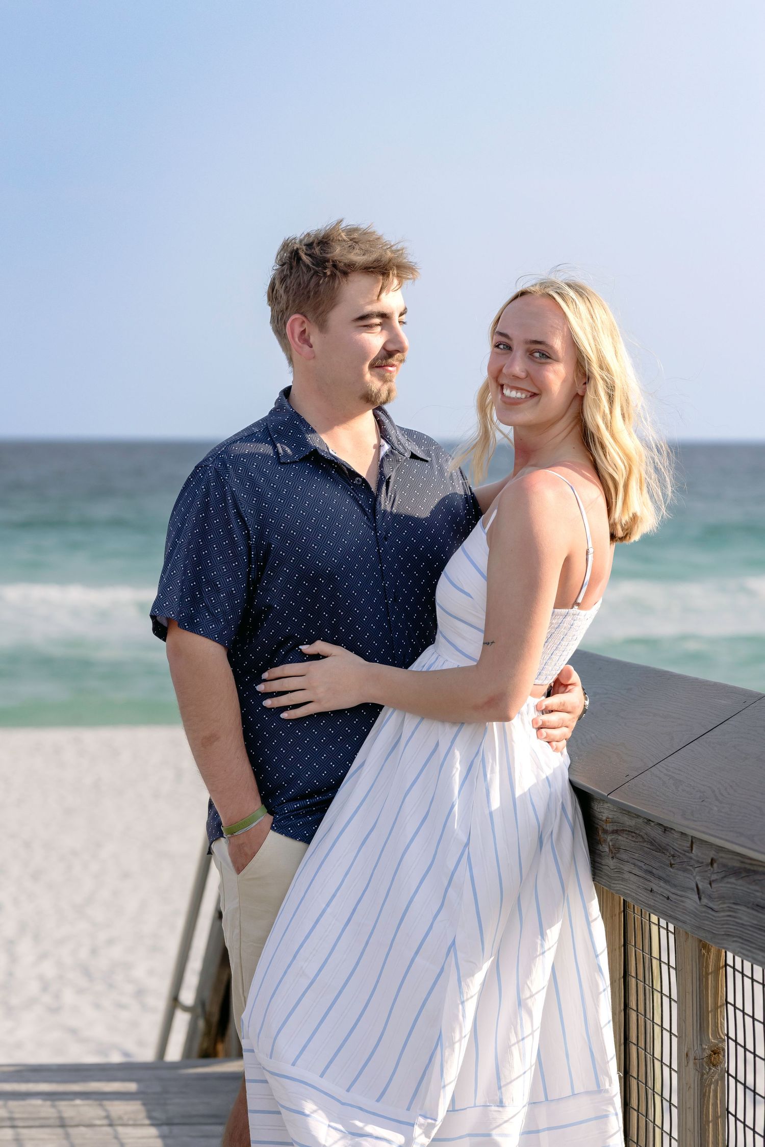 Smiling couple embracing on a sunny beach boardwalk with blue ocean in the background.