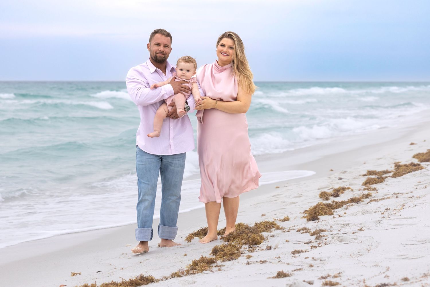 Family of three poses on a sandy beach, parents holding baby, wearing pink outfits with ocean waves behind them.