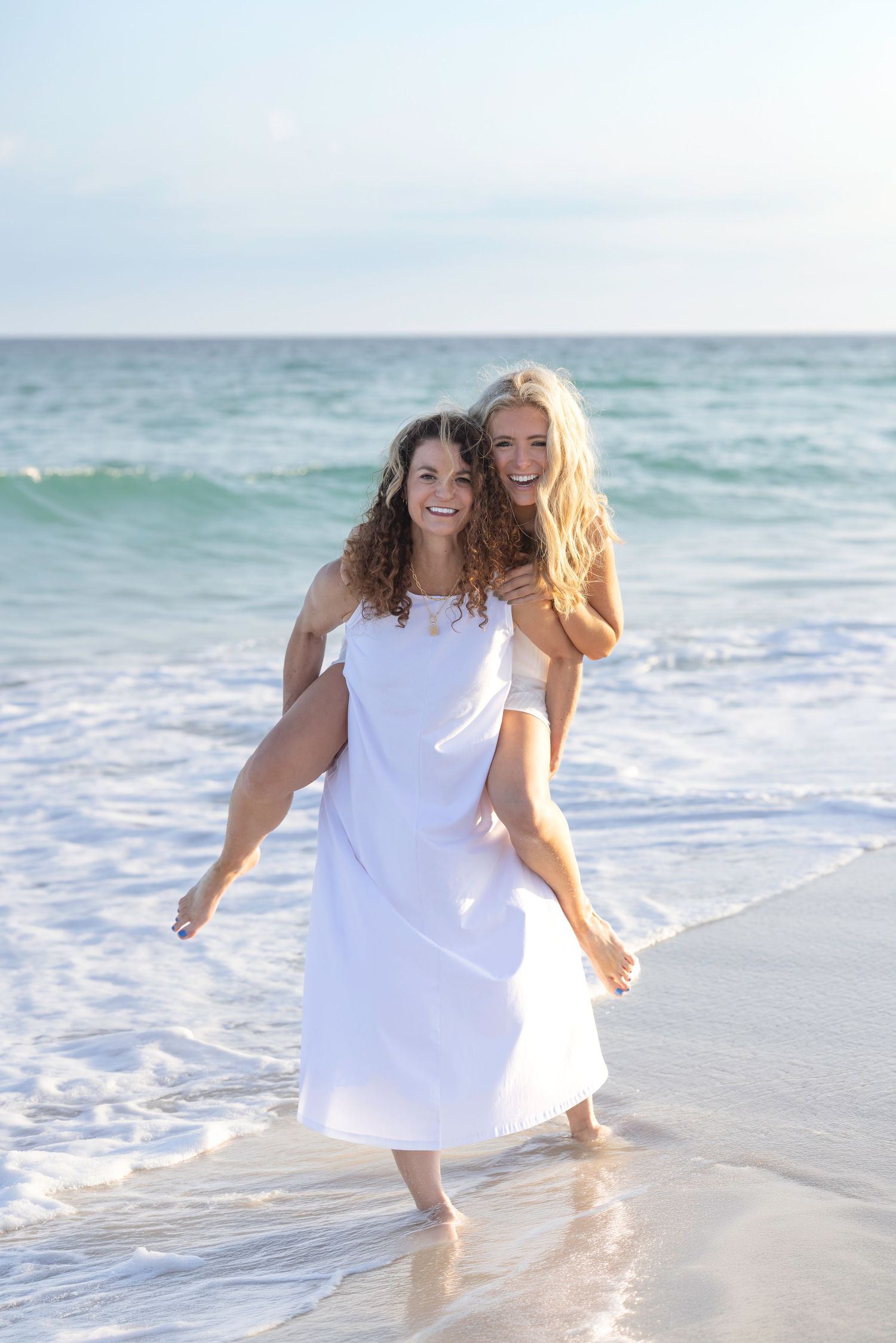 Two women laughing and enjoying a piggyback ride on a sunny beach with ocean waves behind them.