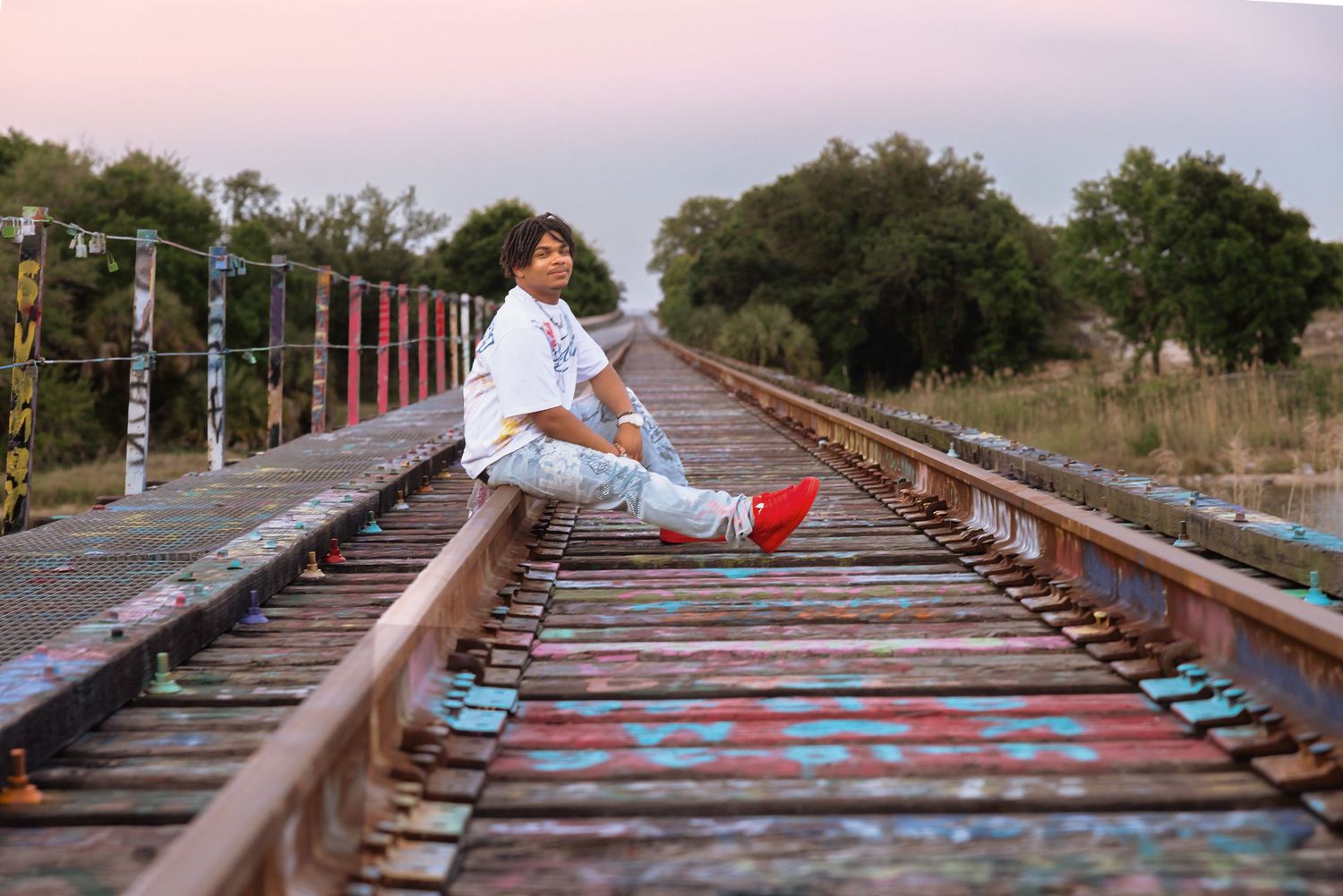 Young man in white shirt and red sneakers sitting on colorful graffiti-covered railway tracks at dusk.